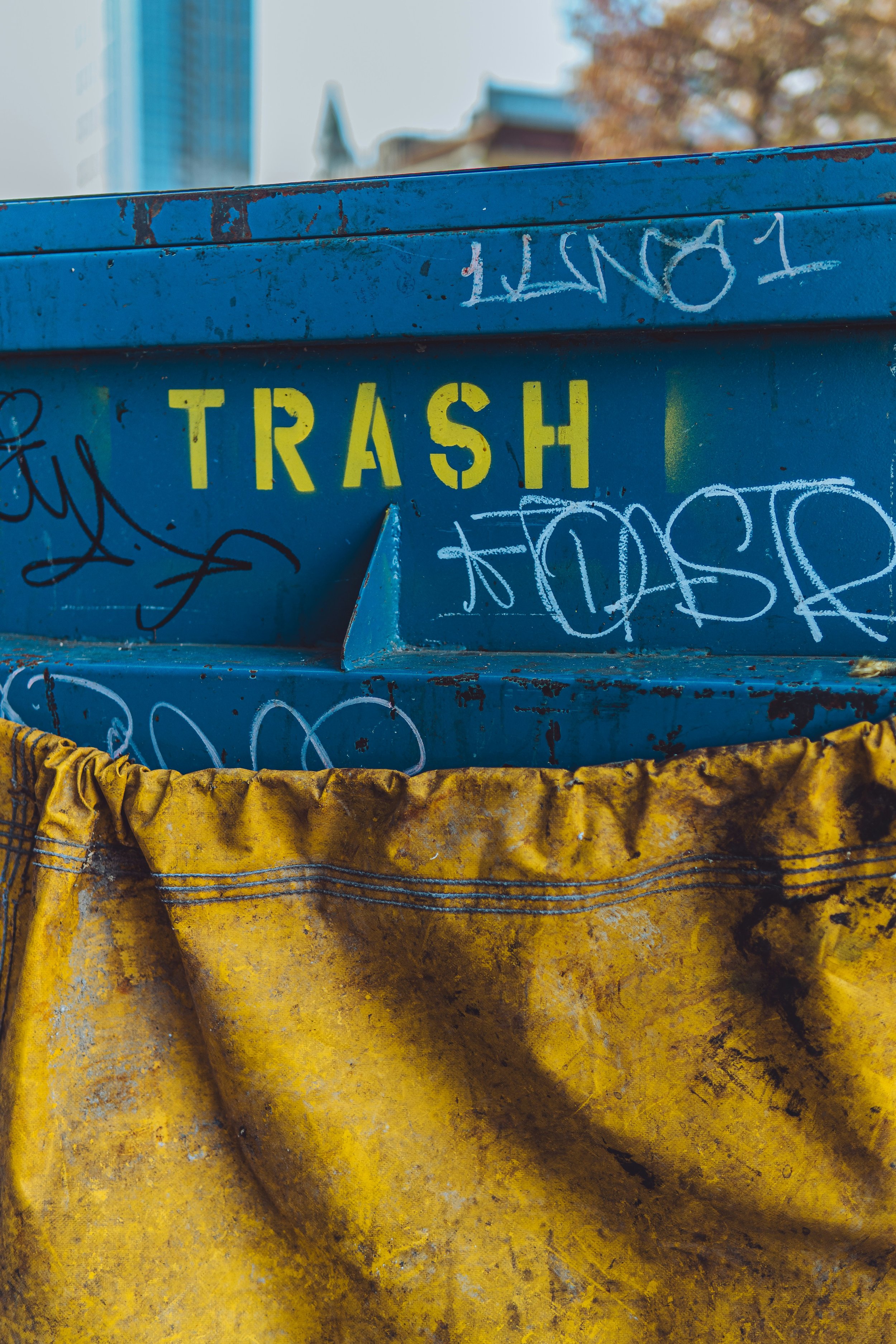 Blue industrial trash container with graffiti, rust, and a yellow fabric covering at the bottom, with trees and buildings in the background.