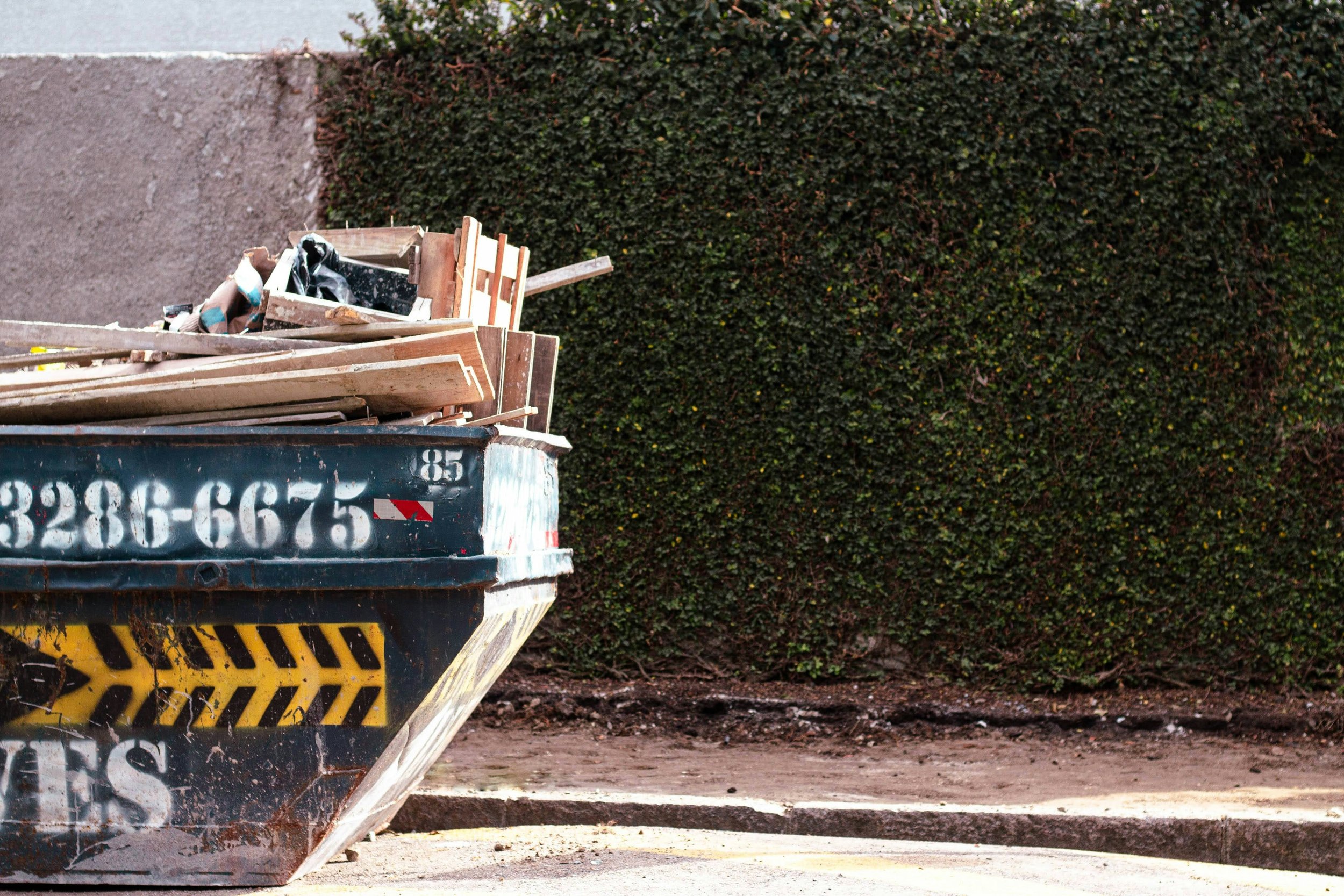 A black dumpster filled with construction debris and wooden planks, set on a street with a tall hedge in the background.