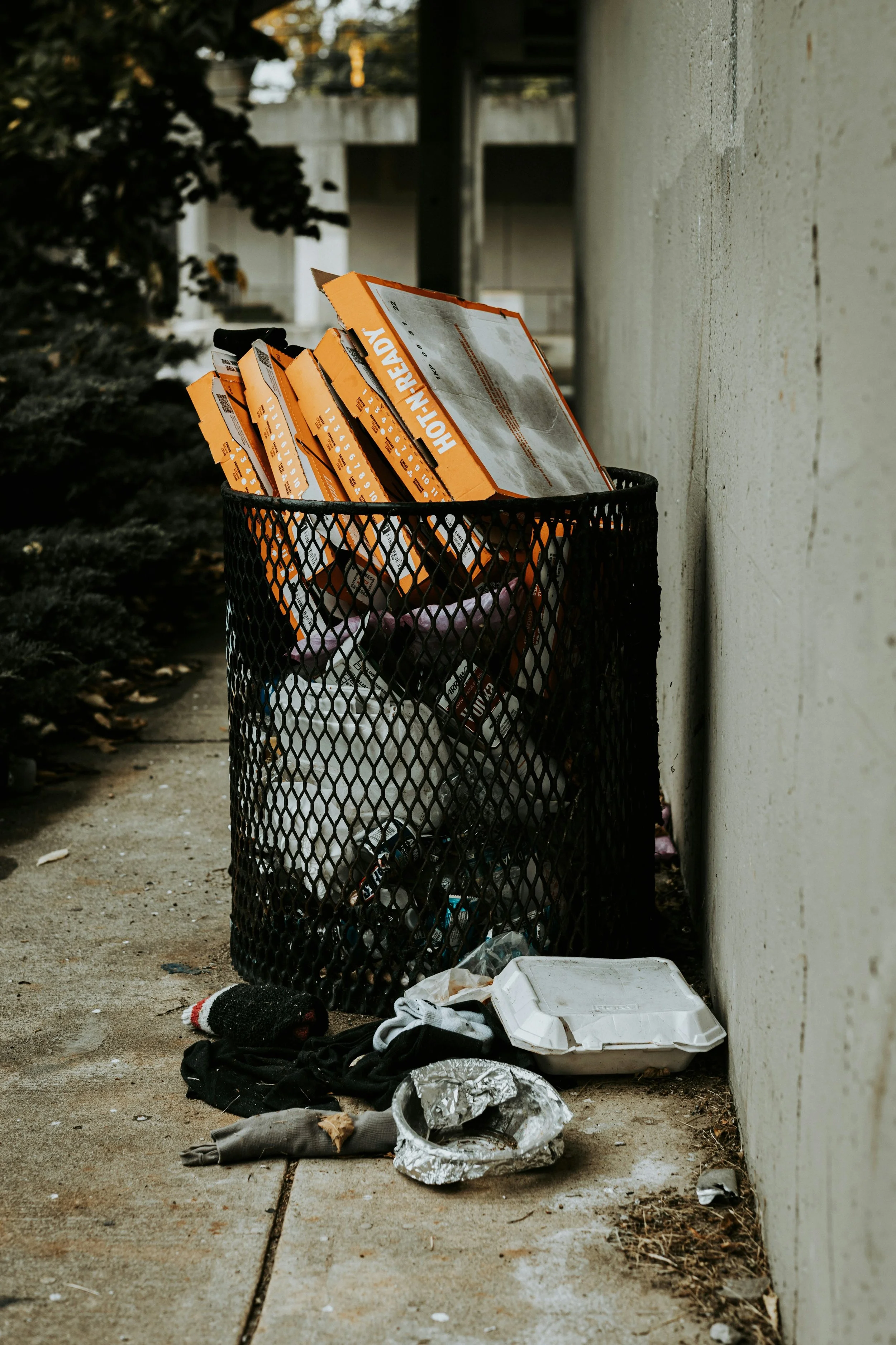 Littered trash and debris around a black metal trash bin, including cardboard pizza boxes, clothes, plastic containers, and other waste, on a concrete sidewalk beside a white wall.