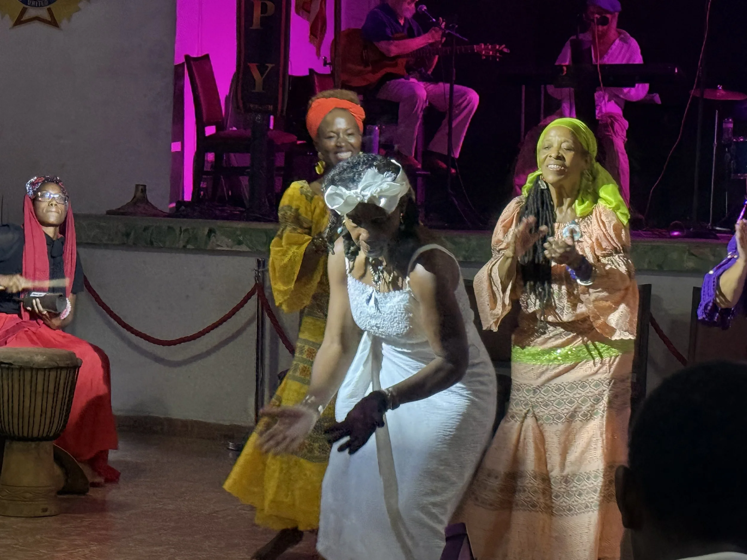 Women wearing traditional African clothing and headwraps dancing on stage, with musicians playing guitars and keyboard in the background, illuminated by colorful stage lights.