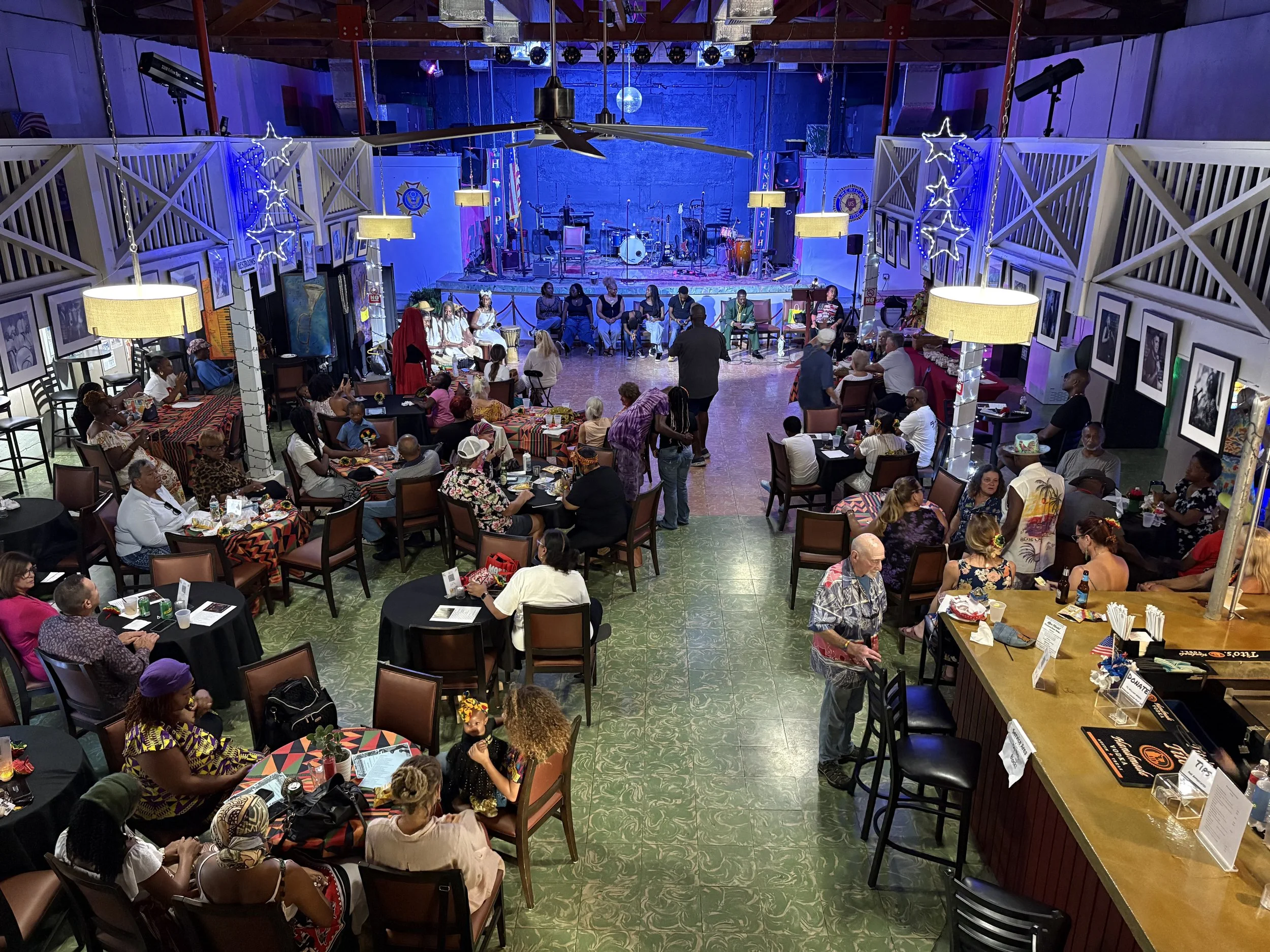 A lively indoor event with people gathered around tables, a stage with musical instruments, and decorative lighting including star-shaped lights and hanging lamps.