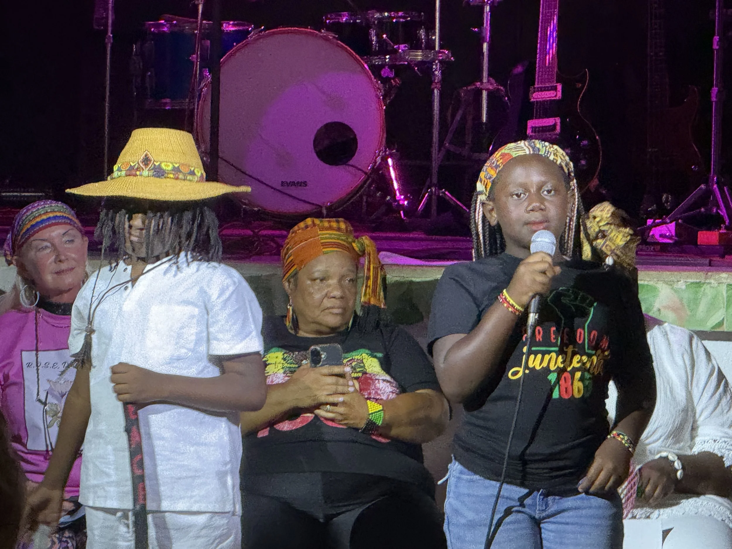 A group of people wearing colorful clothing and accessories, some holding phones, standing in front of a stage with musical instruments and equipment under purple lighting.