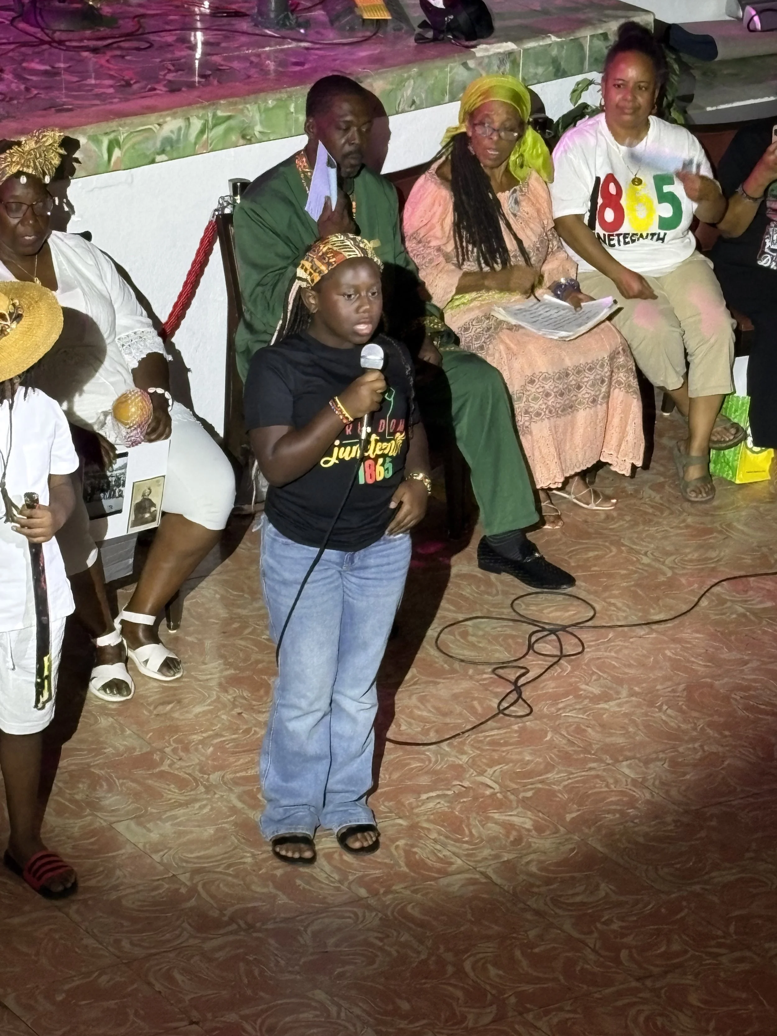 People sitting on chairs, with one woman standing in front holding a microphone, at an indoor event or gathering.