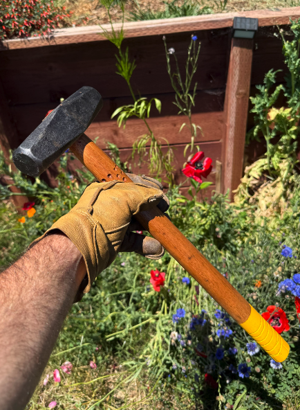 A person's hand wearing a work glove holding a sledge hammer over a flower garden with various colorful flowers and a wooden fence in the background.