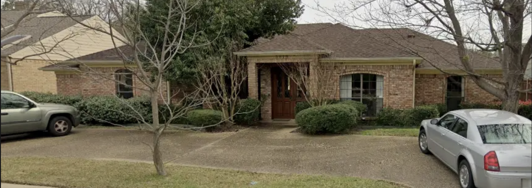 A brick house with a brown roof, surrounded by trees and bushes, two cars parked in the driveway, leafless trees indicating a winter or early spring season.