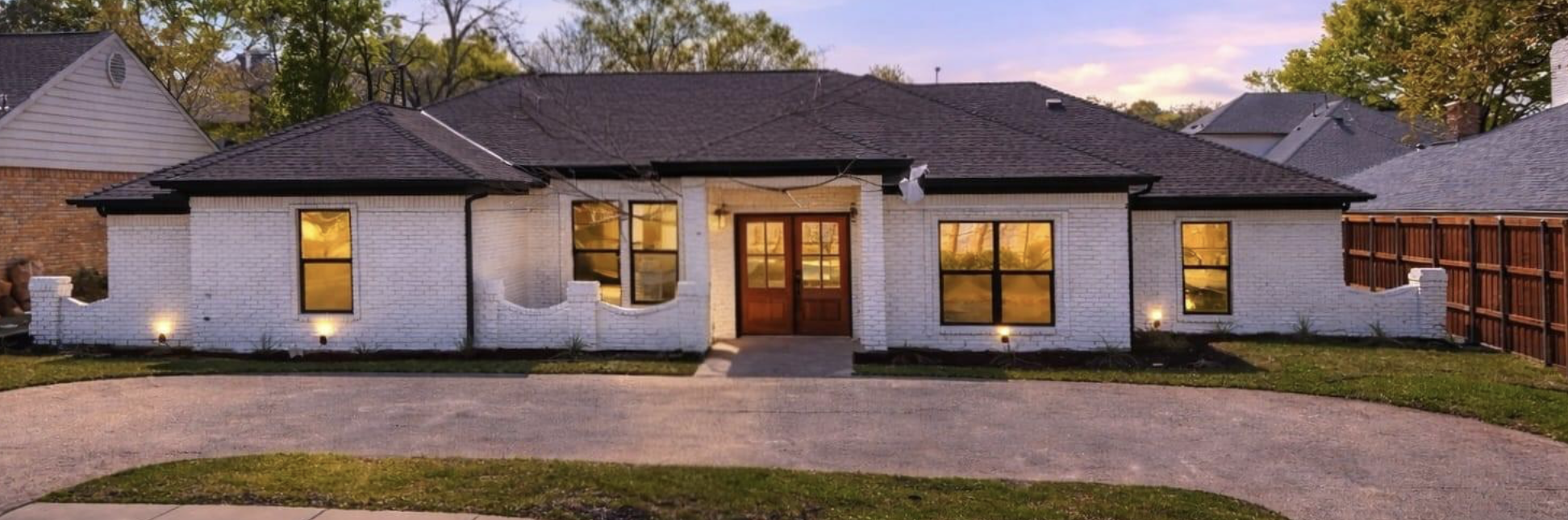 A house with white brick exterior, black window frames, and a wooden door, illuminated by exterior lights at dusk, with a driveway and a wooden fence on the right.