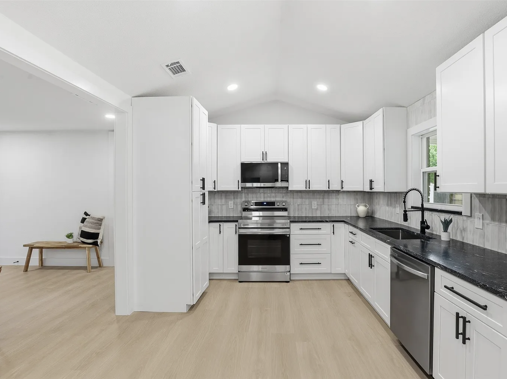 Modern white kitchen with black countertops, stainless steel appliances, black cabinet handles, a black sink with a black faucet, a window with greenery outside, and small decorative items on the counter. Adjacent room features a wooden bench with a plant, pillow, and wall art.