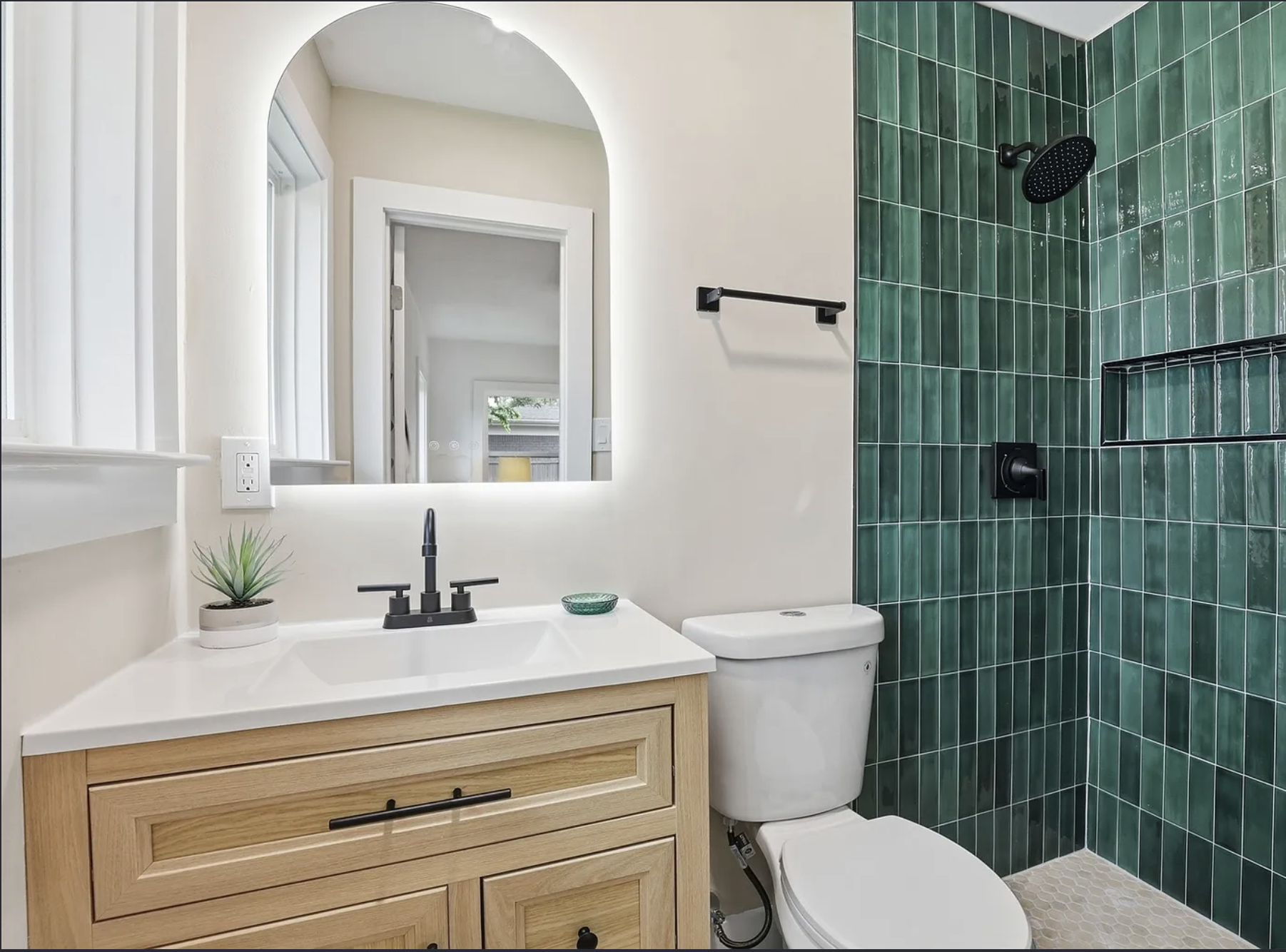 Bathroom with a wooden vanity, white sink, black faucet, black mirror, small potted plant, toilet, and green tiled shower area with black fixtures.