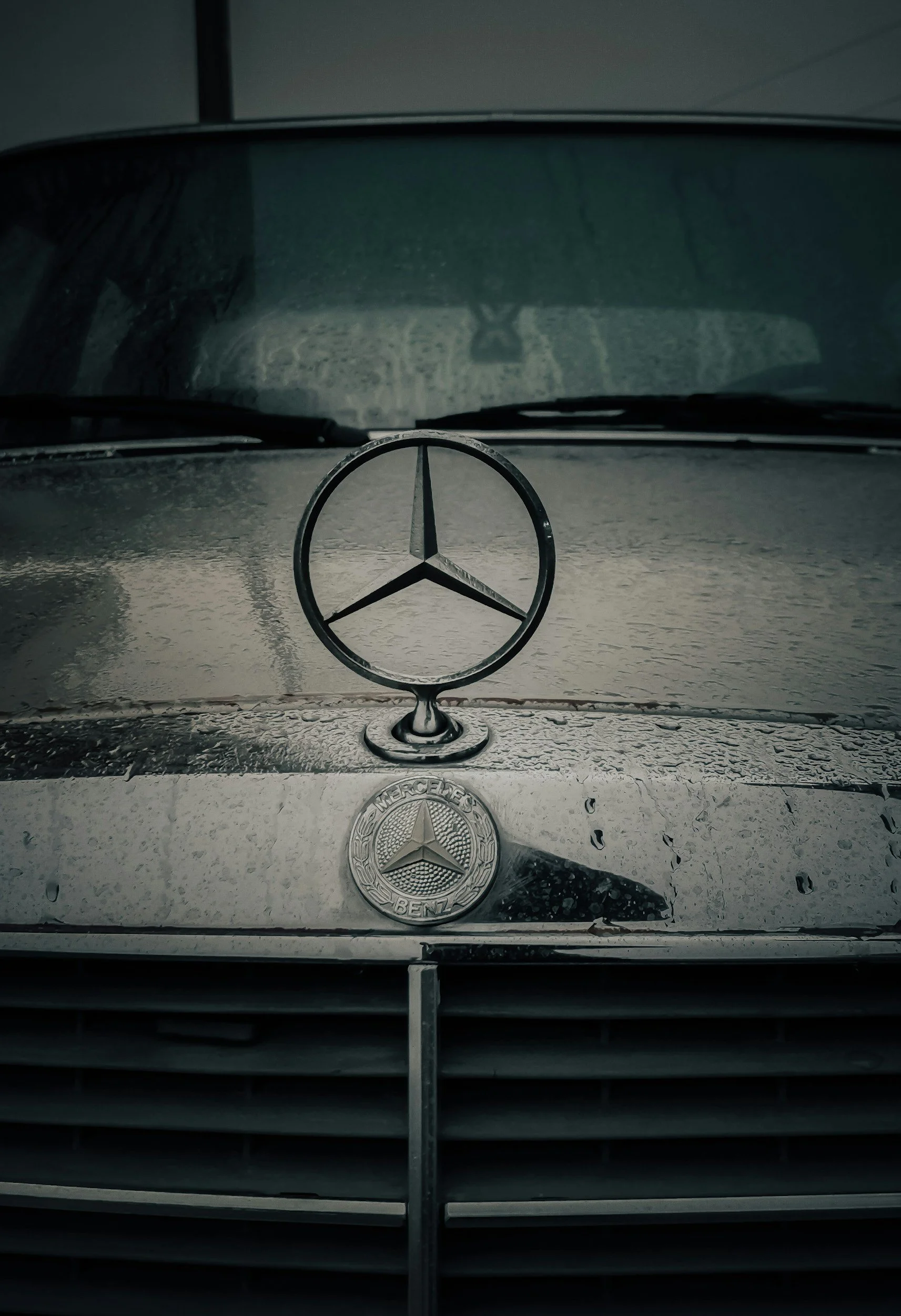 Front view of a Mercedes-Benz car with the hood ornament and grille visible, rain on the windshield, and the Mercedes-Benz emblem on the hood.