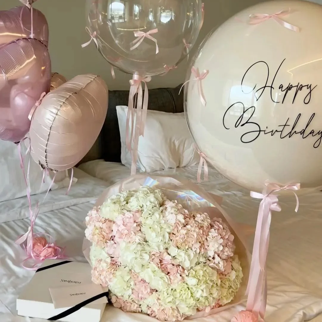 A bed decorated with pink and clear balloons, a bouquet of pink and white hydrangeas, and a birthday message on a clear balloon. The scene suggests a celebration, likely a birthday.
