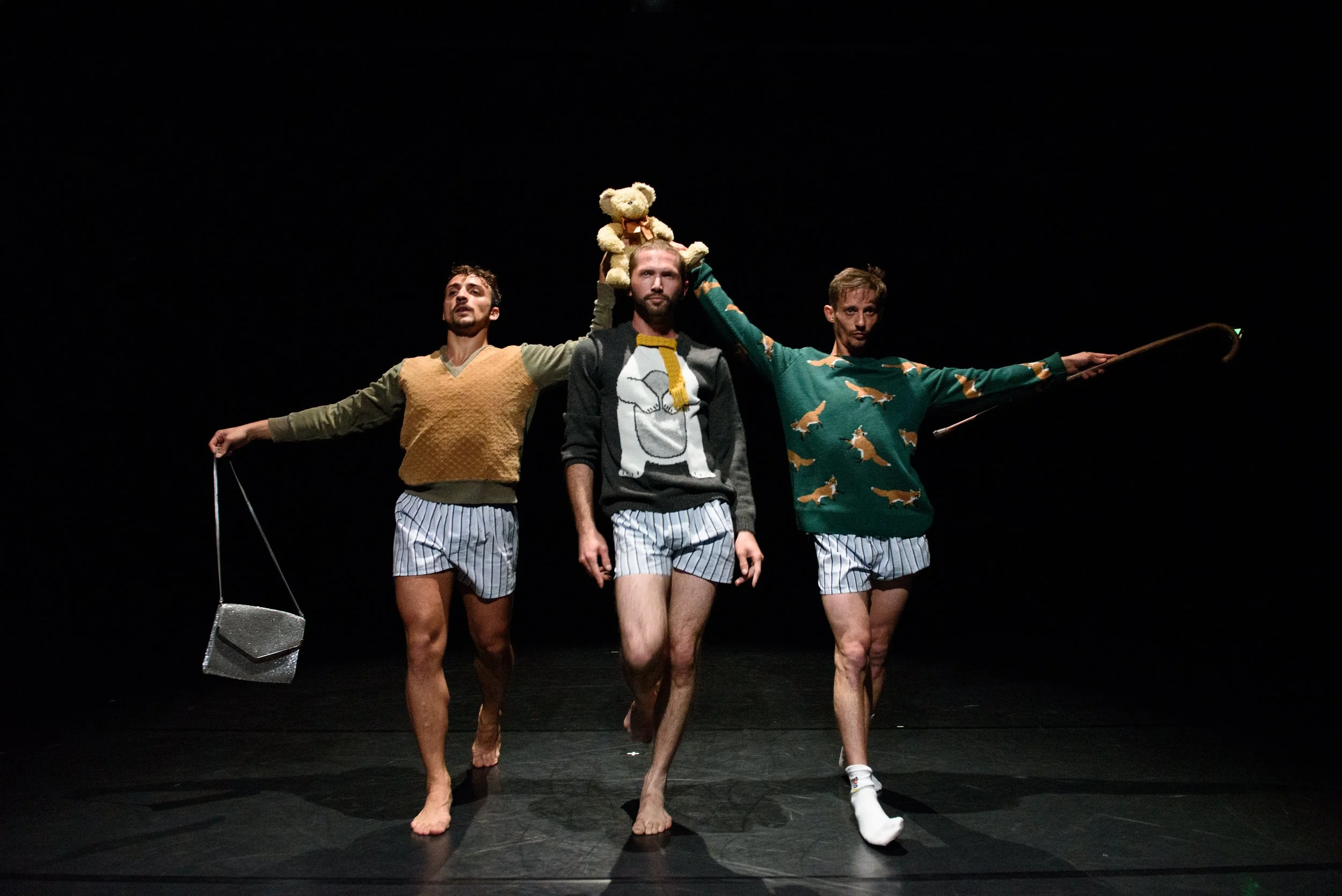 Three men walking barefoot on stage, wearing pajama shorts and sweaters, with a black background. The man to the right holds a shepherd's staff, the man in the center carries a teddy bear on his head, and the man to the left holds a small box.
