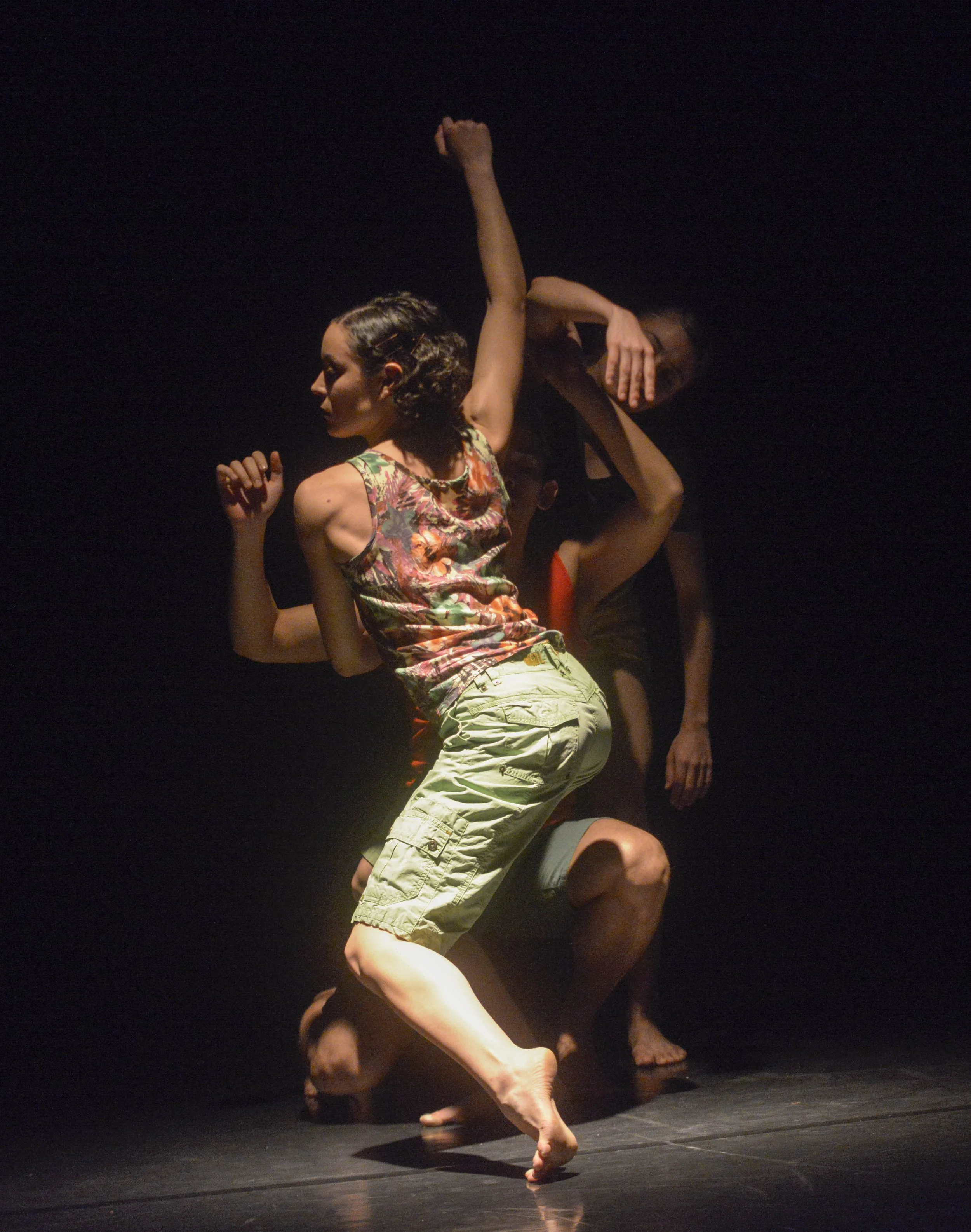 Dancers performing on stage with black background, one dancer lifting another, wearing colorful and beige shorts, with focused expressions.
