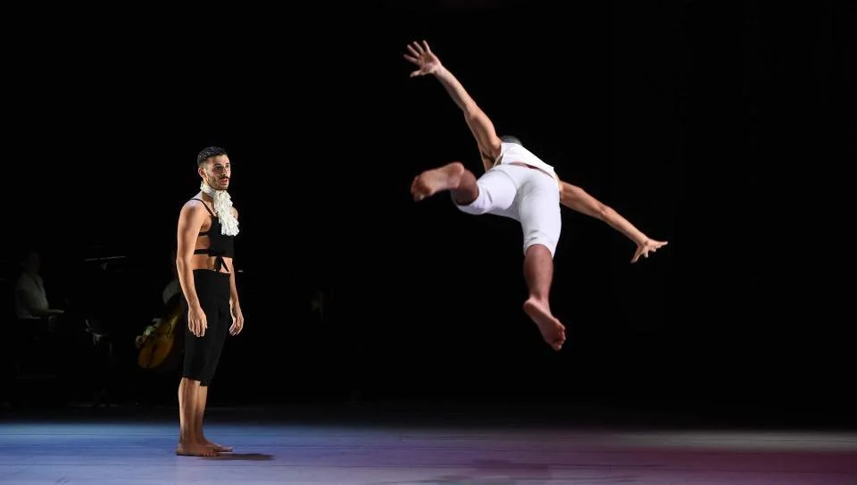 A male dancer performing a flip in mid-air during a dance performance, with a male dancer standing on the stage watching.