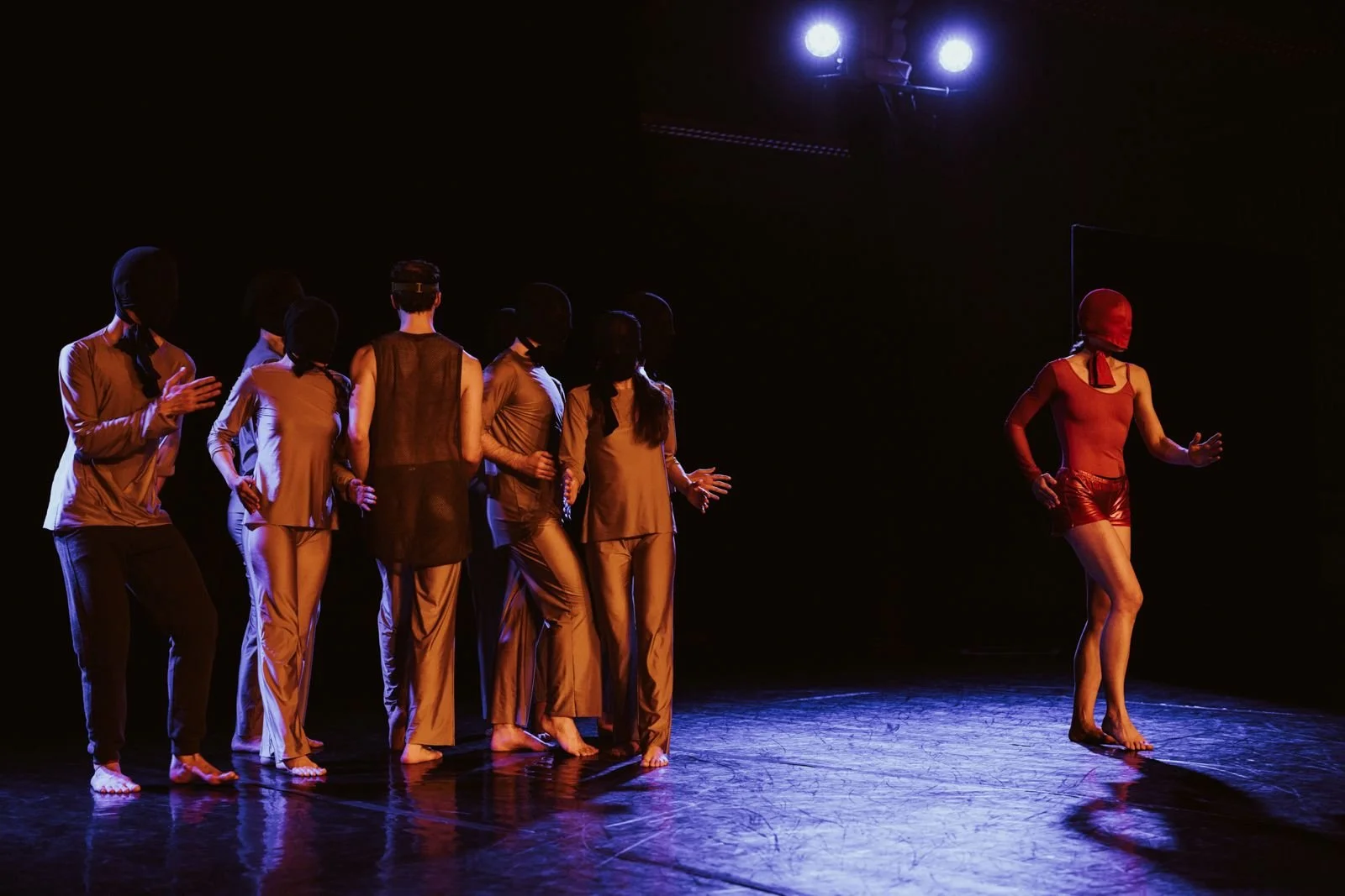 A group of seven dancers on stage, wearing beige and black attire with black masks, facing left toward a dancer in red who is also masked and is barefoot, with dark background and stage lights overhead.