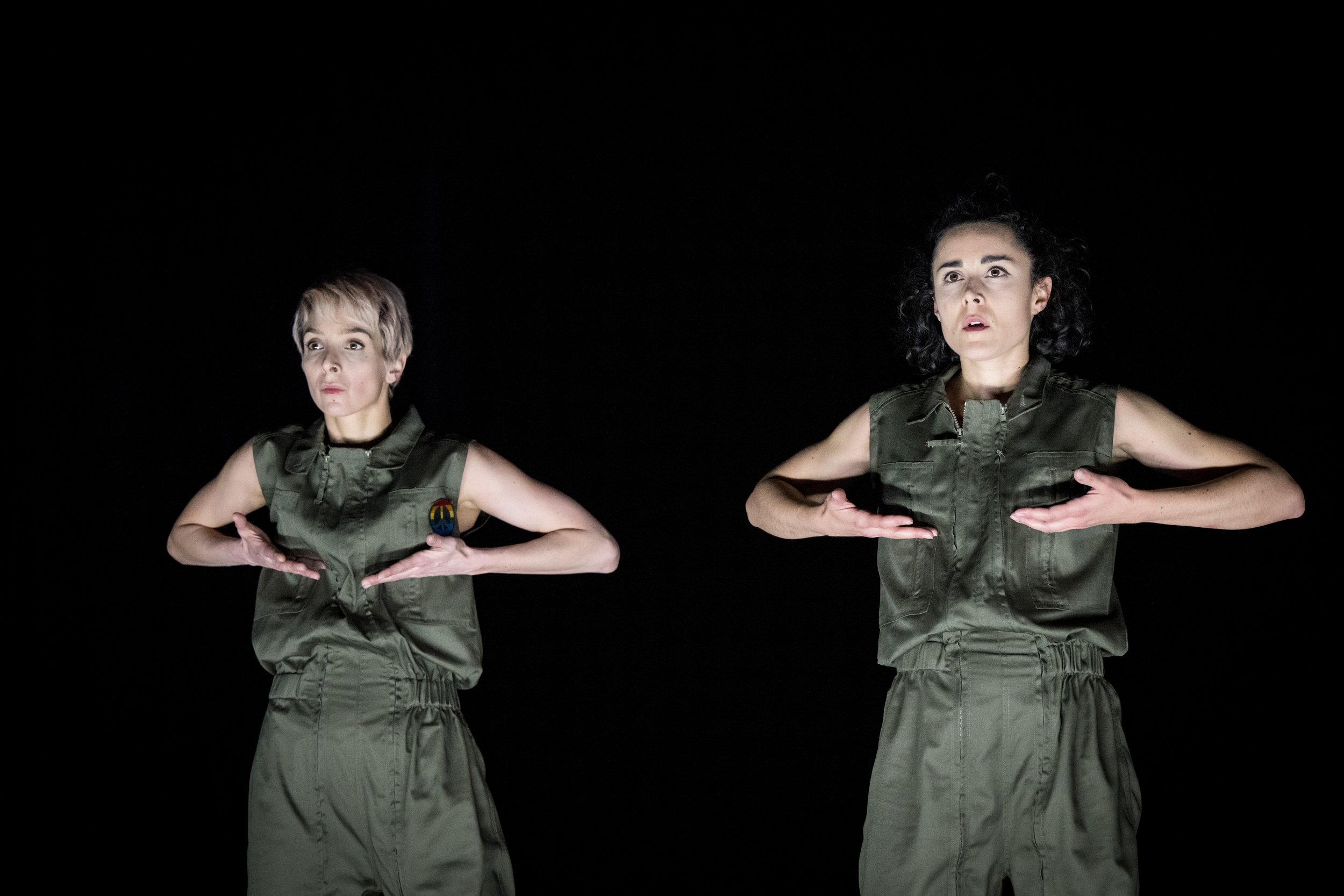 Two women with dark curly and blond straight hair, respectively, dressed in khaki military-style outfits, perform a dance move with their hands on their hips against a black background.