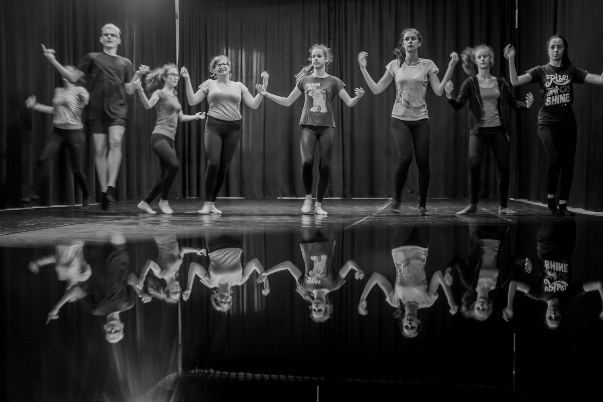 Group of children and a young adult holding hands and dancing on a stage with a black curtain backdrop.