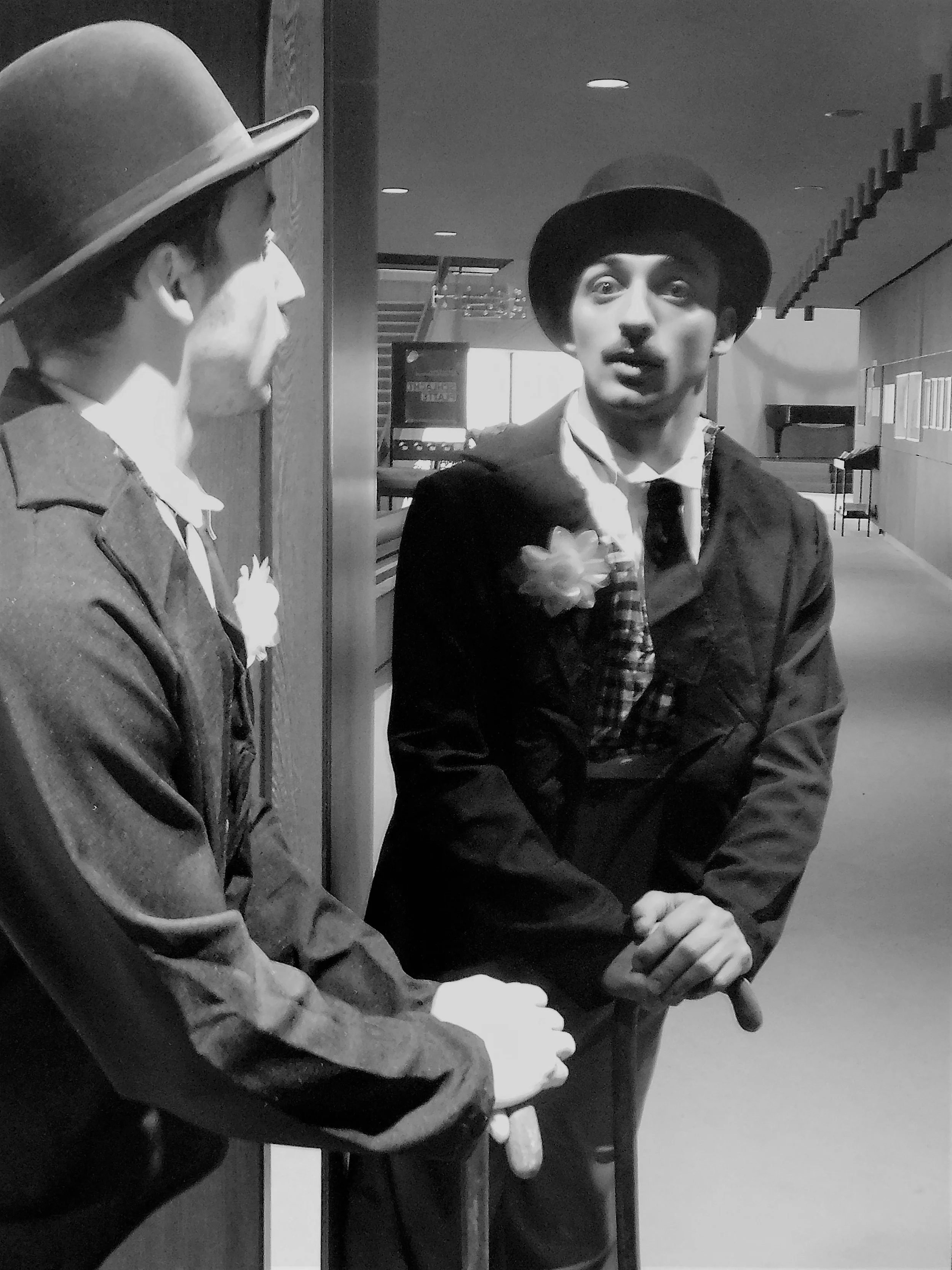 Two men wearing suits and bowler hats standing and talking to each other in an indoor setting.