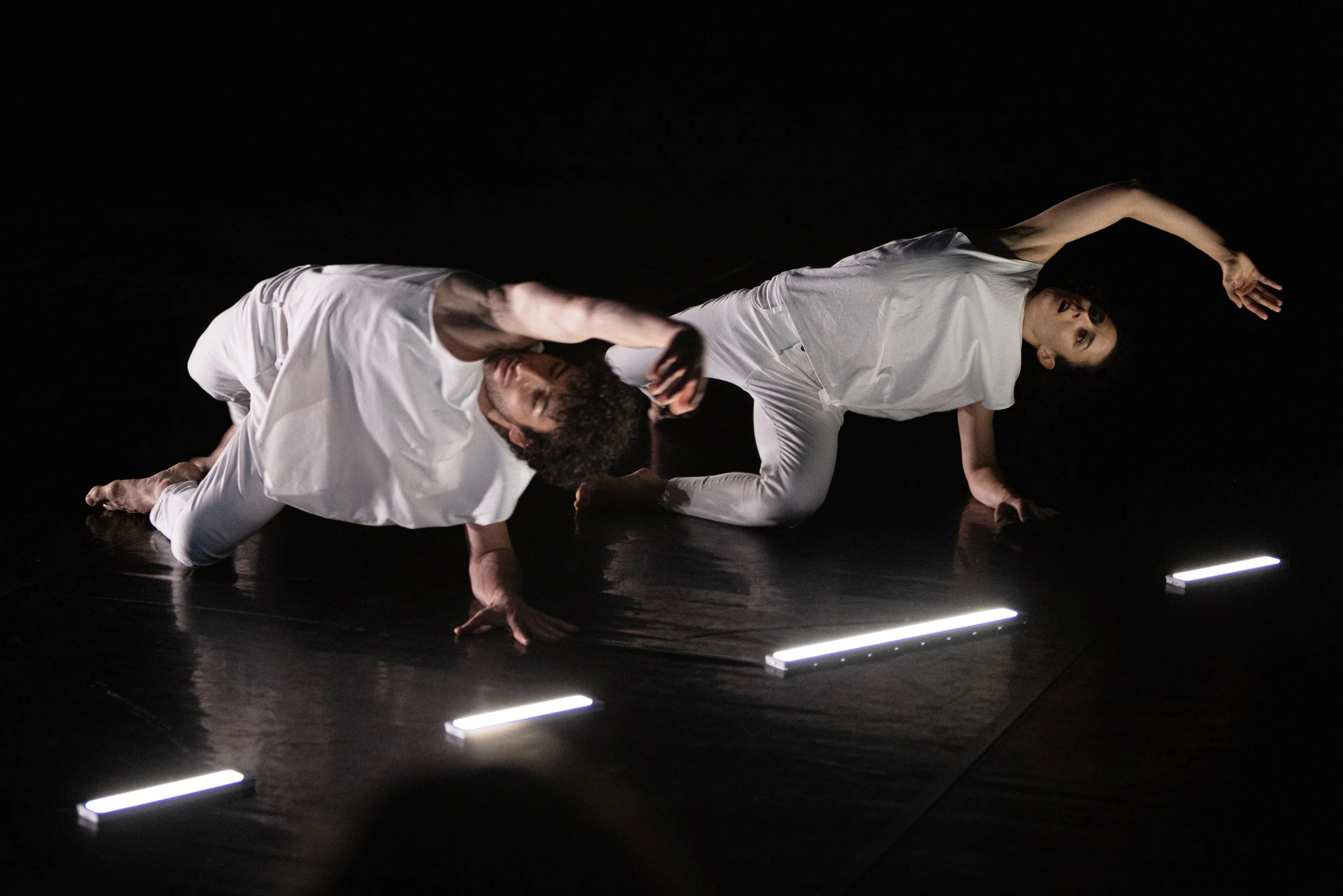 Two dancers in white outfits perform on a dark stage with minimal lighting.