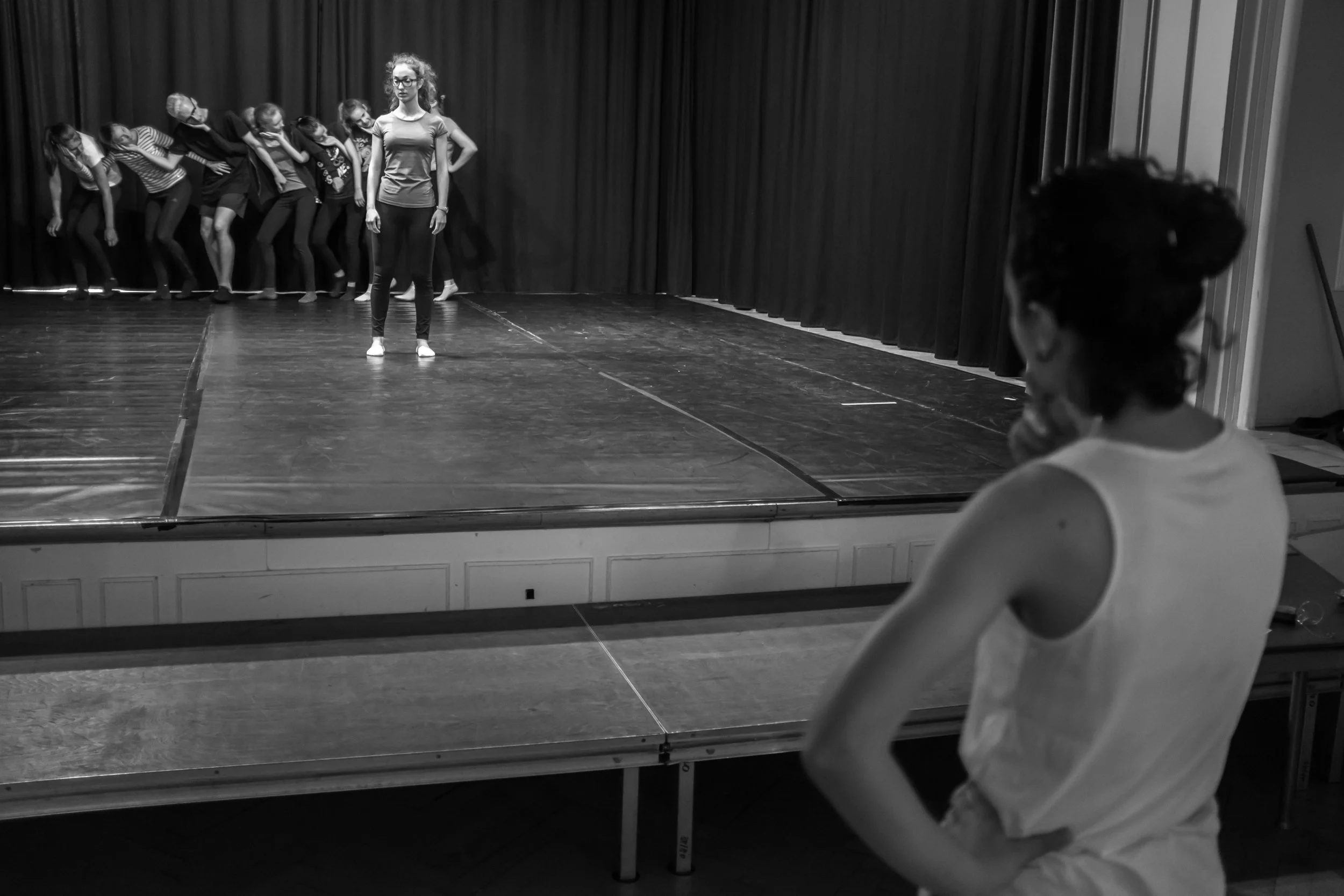Black and white photo of a stage with a curtain backdrop, featuring the teacher in the center mentoring her Jugendclub tanz.