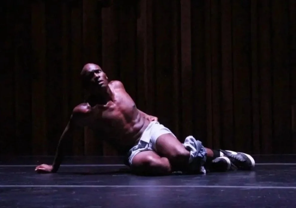 A shirtless man wearing shorts and sneakers is performing on a dark stage with a wooden backdrop, kneeling with one arm supporting him and the other raised.
