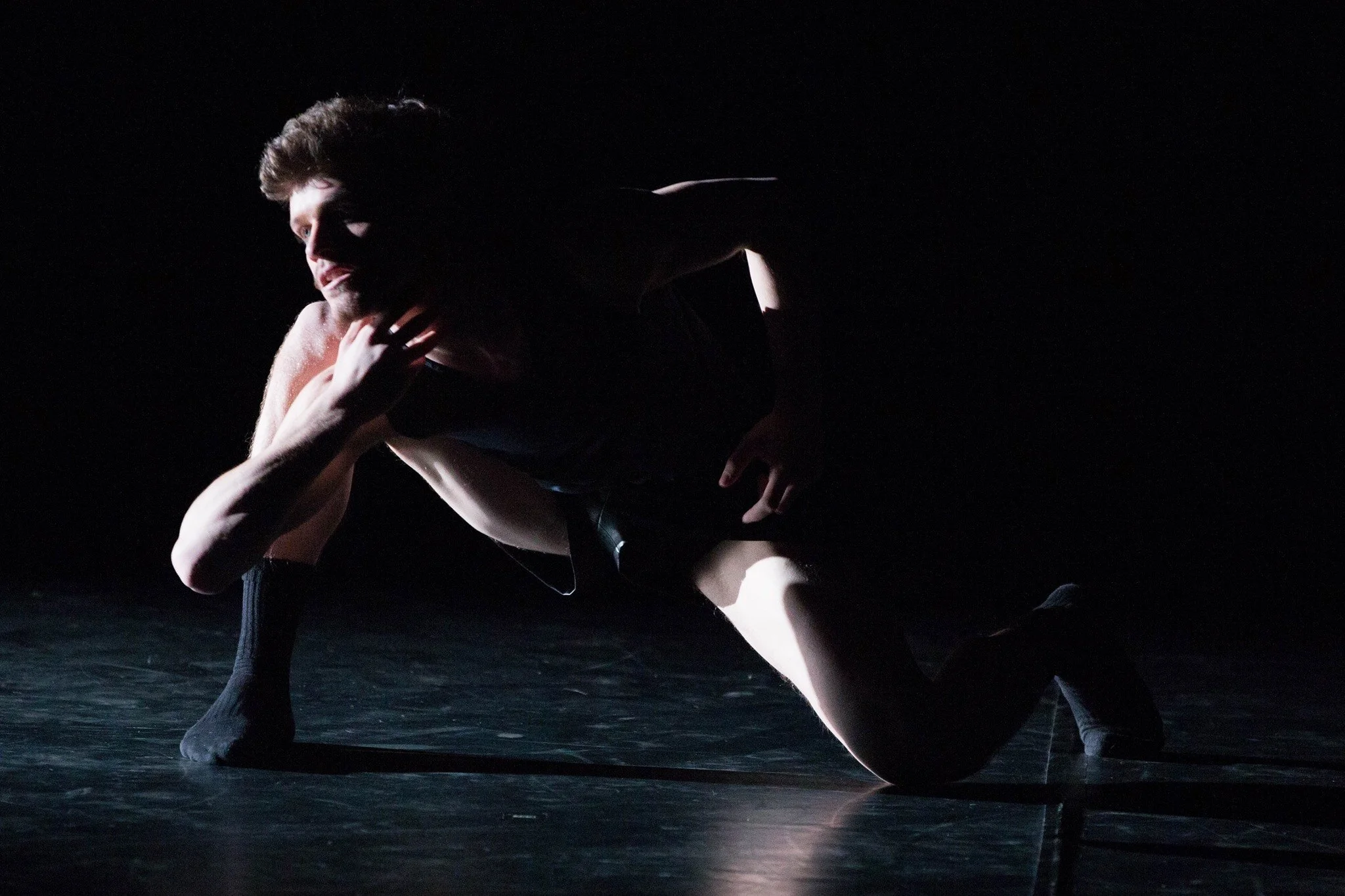 A male dancer performing on a dark stage, illuminated by a focused light, wearing black clothing, in a crouched position with one arm extended forward and the other bent near his face.