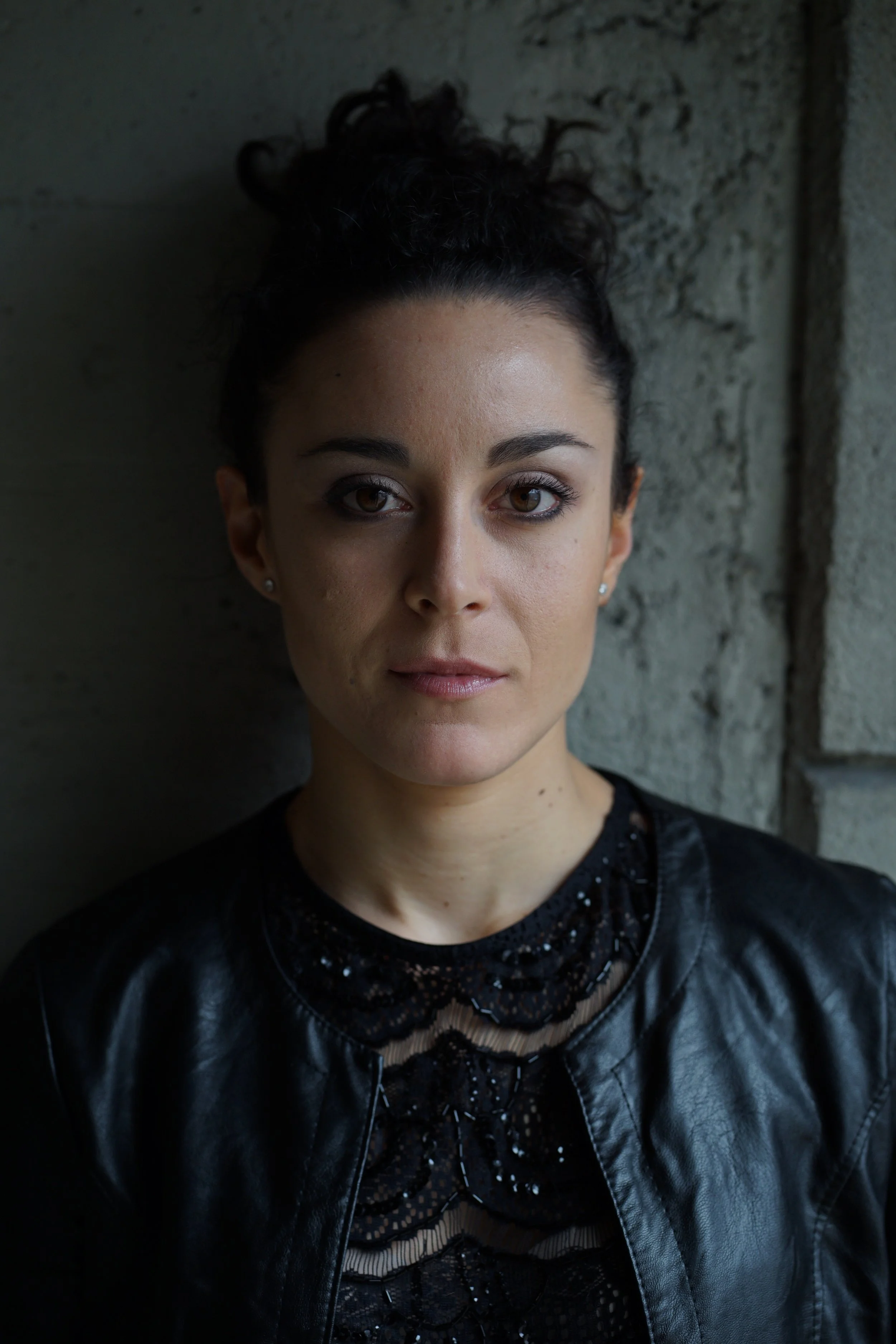 Close-up of a woman with dark curly hair tied up in a bun, standing against a textured gray wall, wearing a black top with lace detail and a leather jacket.