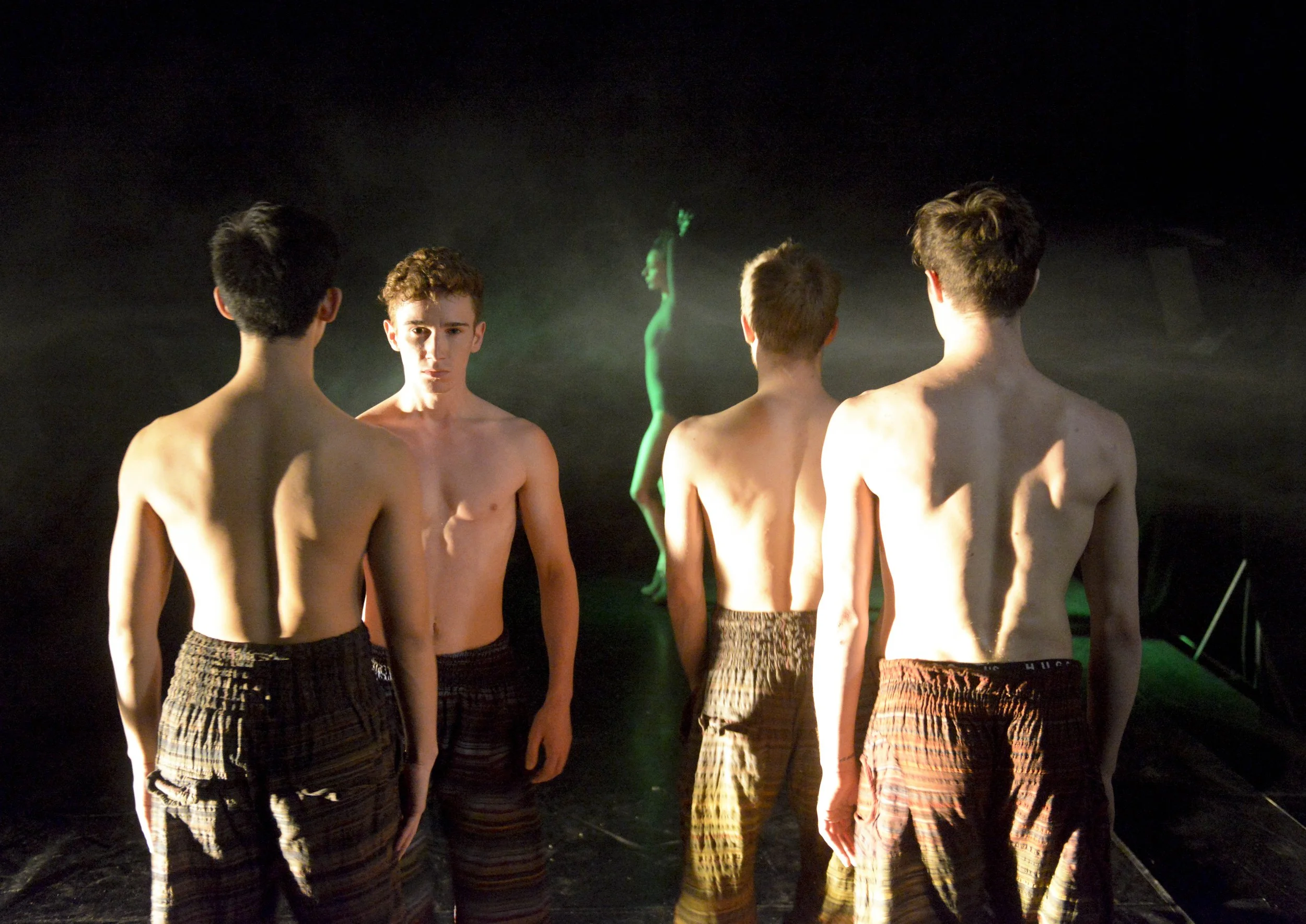 Four shirtless young men with striped shorts standing in a row on a dark stage with a dancer in the background illuminated by green and purple lights.