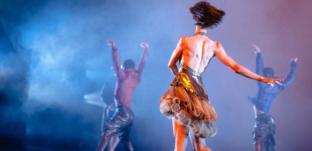A woman dancing on stage with backup dancers, surrounded by colorful lighting and smoke effects.