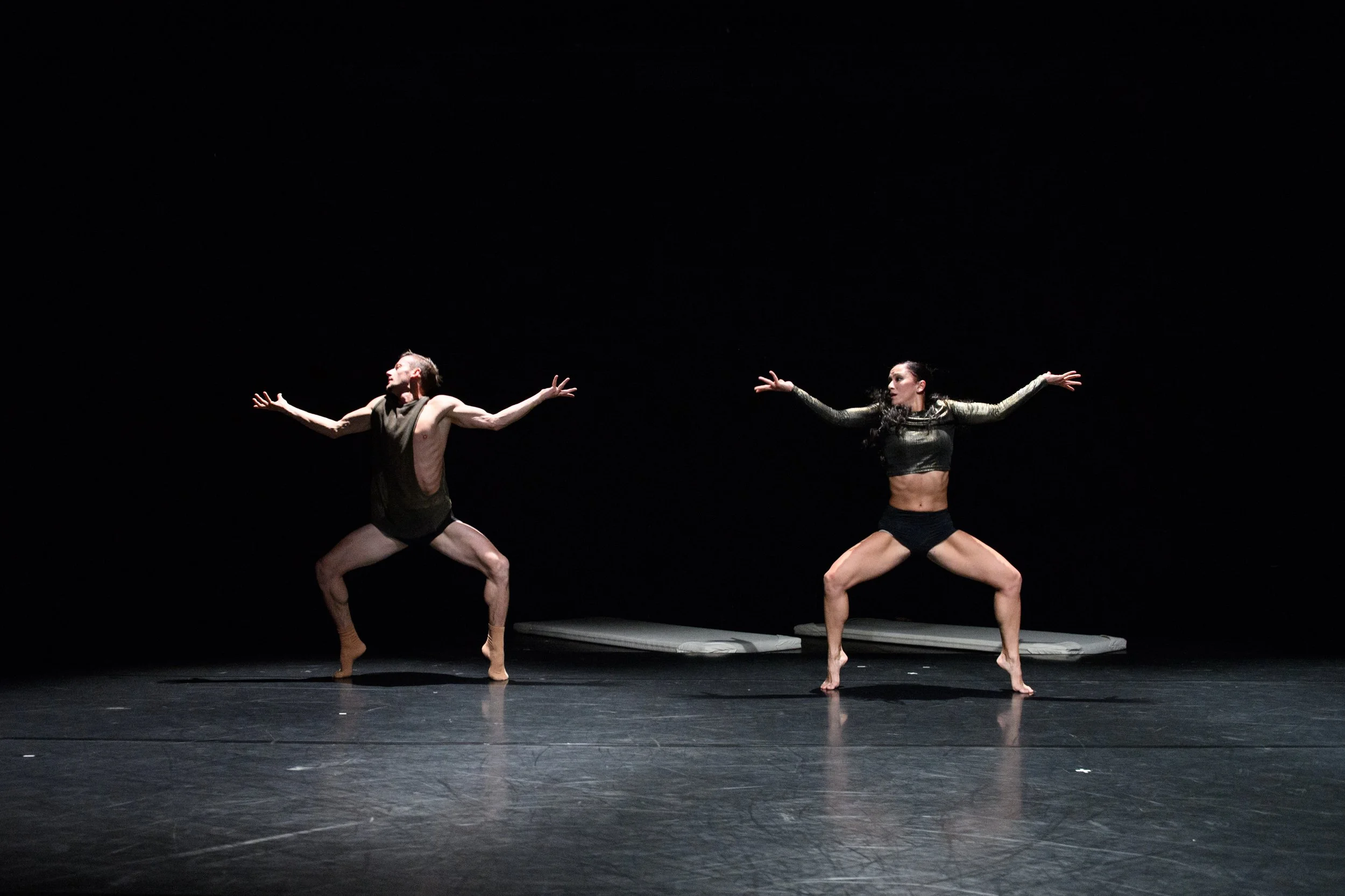 Two dancers performing a contemporary dance routine on stage with a dark background. They are in a wide stance with arms extended, wearing minimal, black costumes.