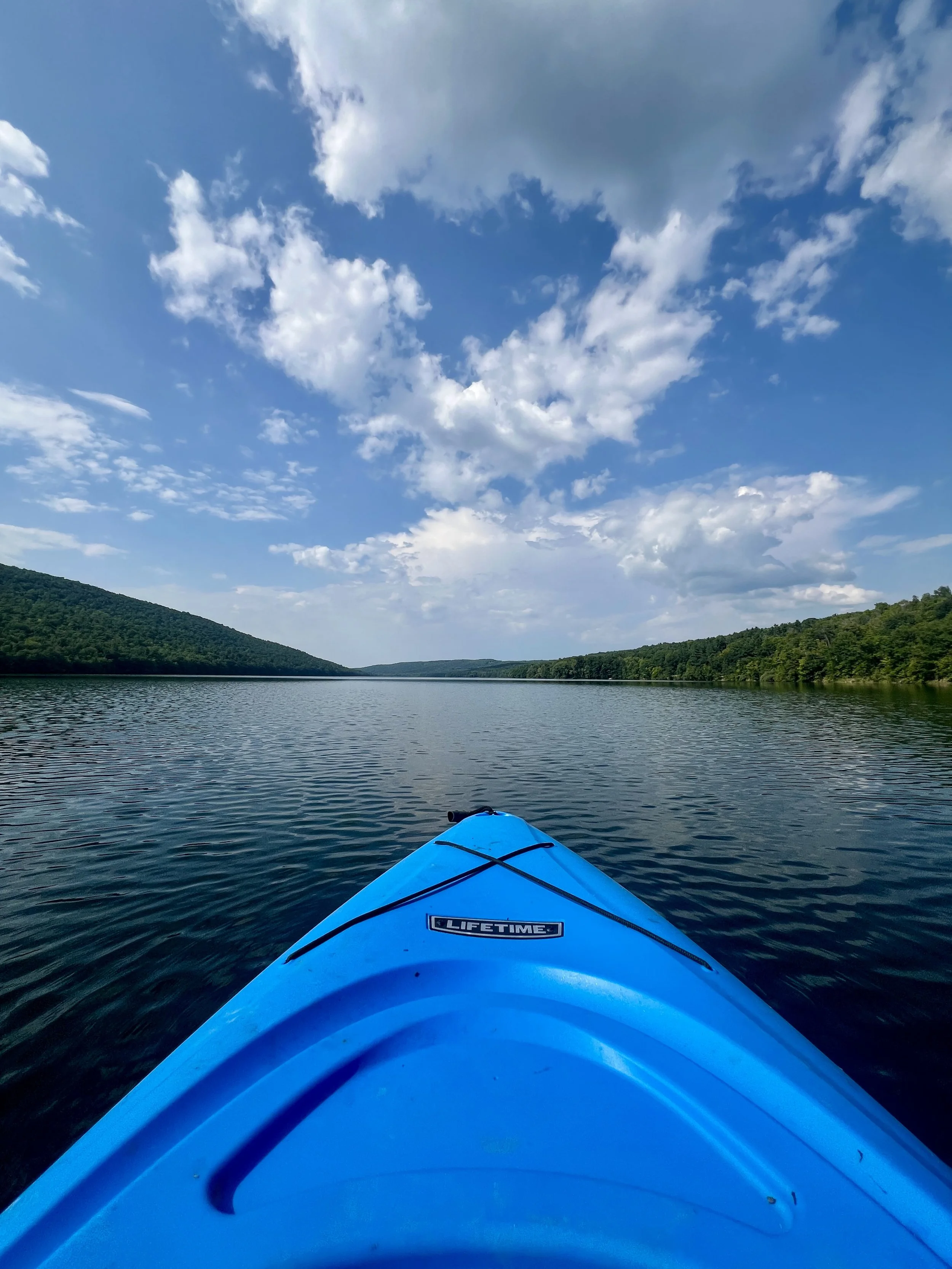 View from a blue kayak on a calm river, with hills covered in trees on both sides and a partly cloudy sky overhead.