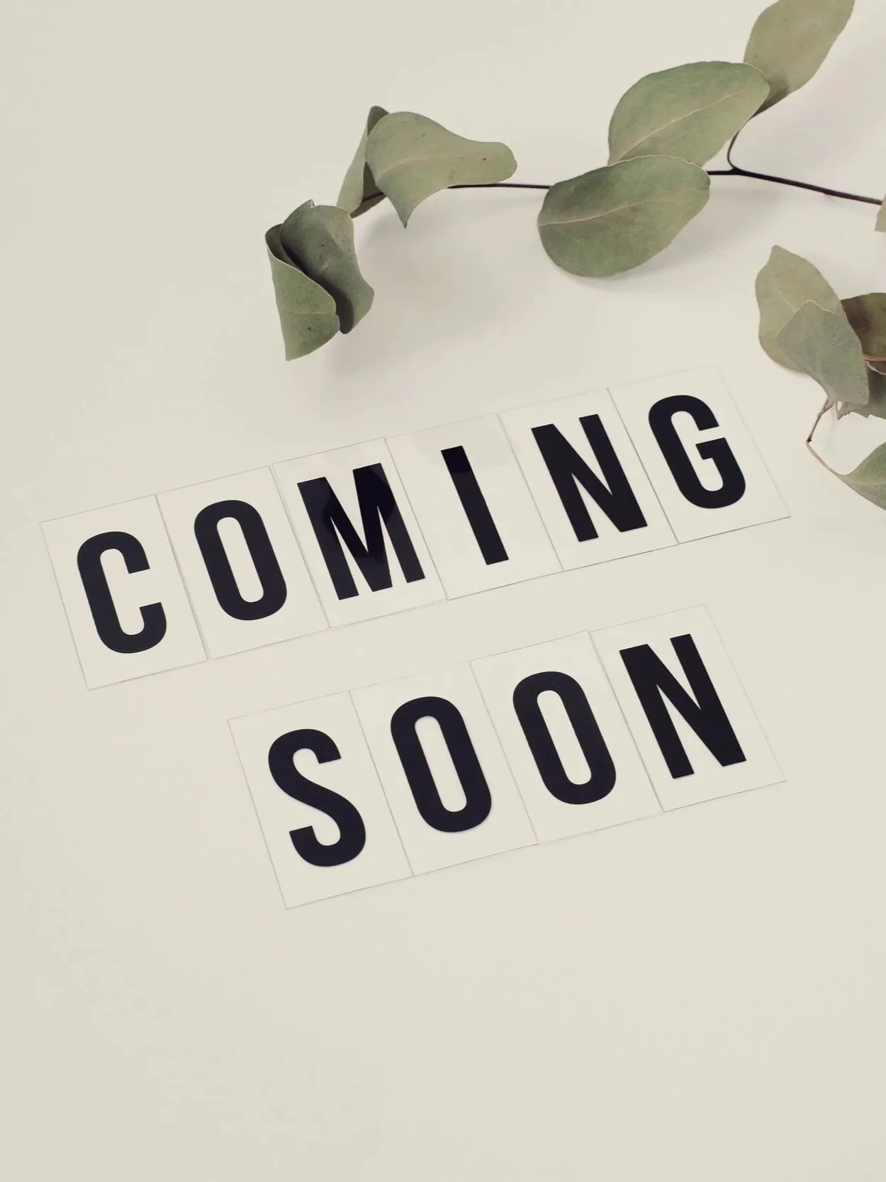 A white surface with black letter tiles spelling out "COMING SOON" and some green eucalyptus leaves placed on top.