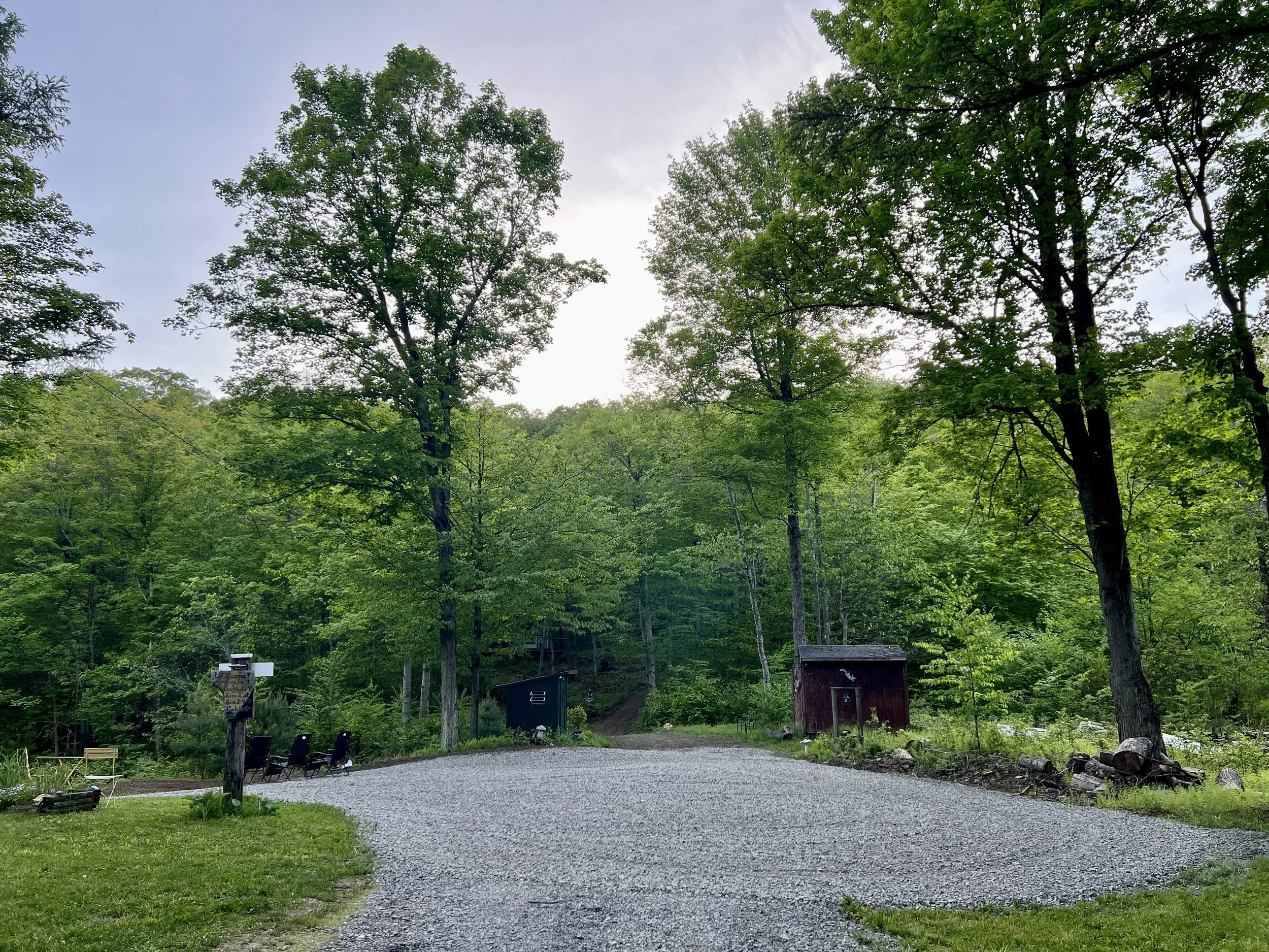 A gravel pathway leading into a wooded area with green trees, two small sheds, and a few outdoor chairs and items near the bottom of the image.