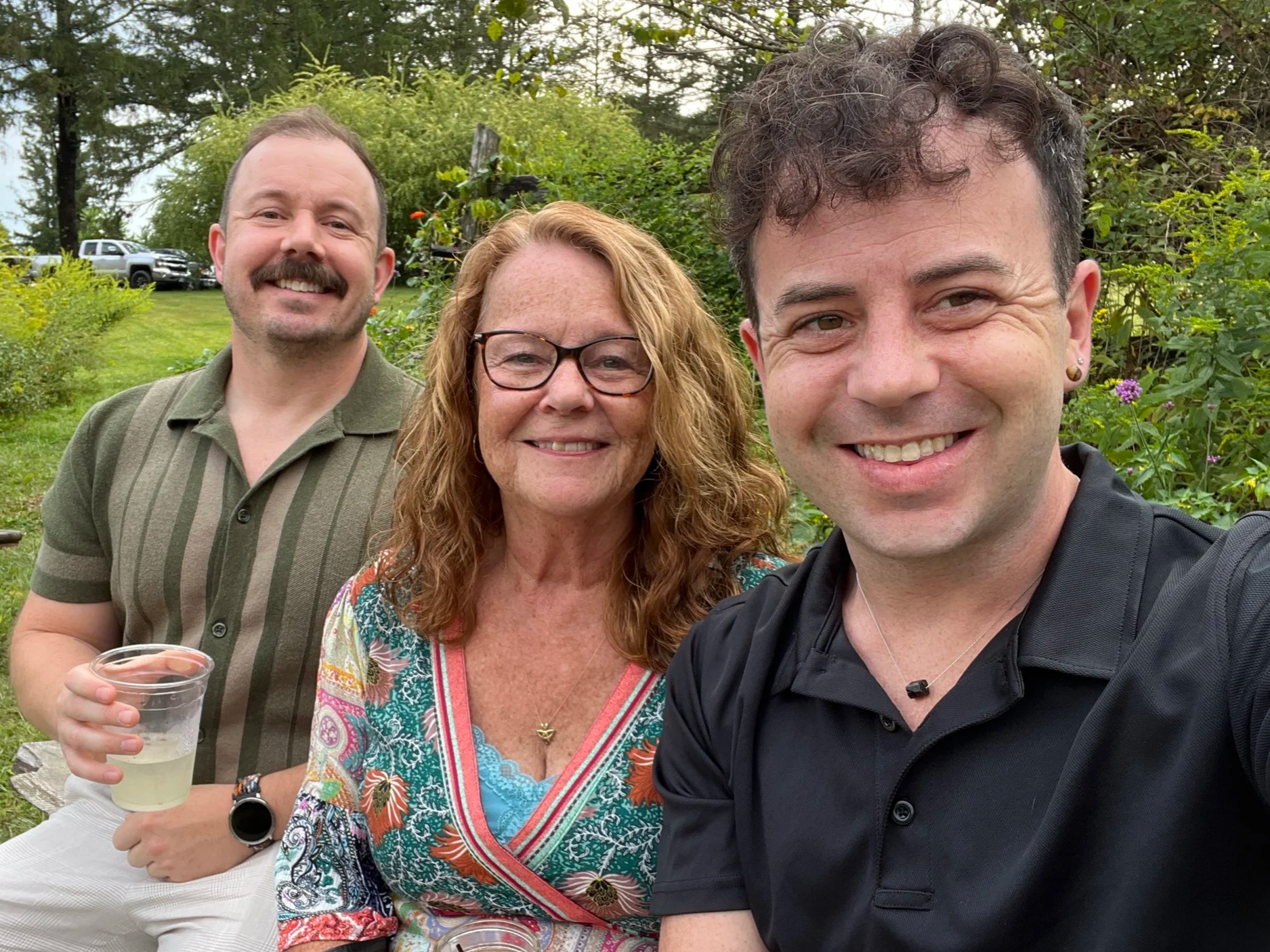 Three people smiling outdoors, with two men and one woman in the center, holding standing in a lush green garden.
