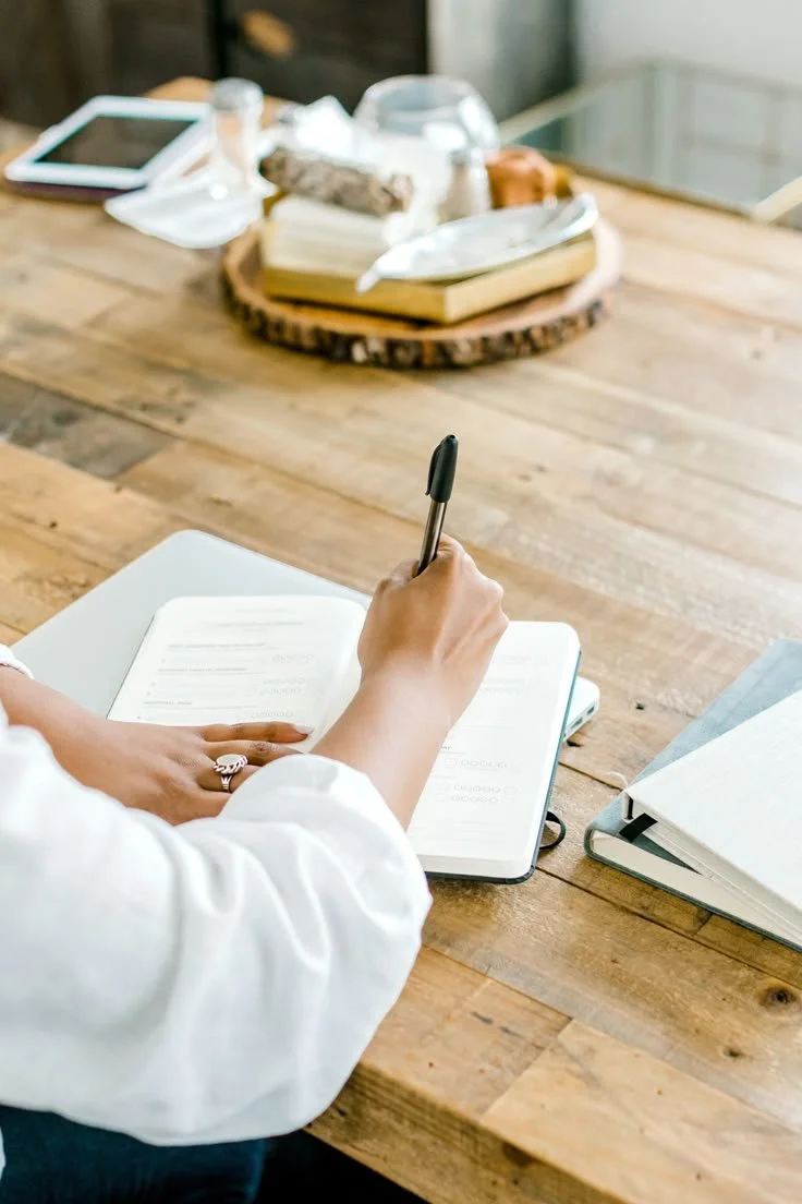 Person writing in a notebook at a wooden table with electronic devices and a snack tray in the background.