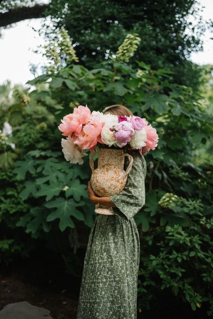 A person in a green patterned dress holding a large beige and brown vase filled with pink and white peonies. They are standing outdoors in front of lush green foliage.