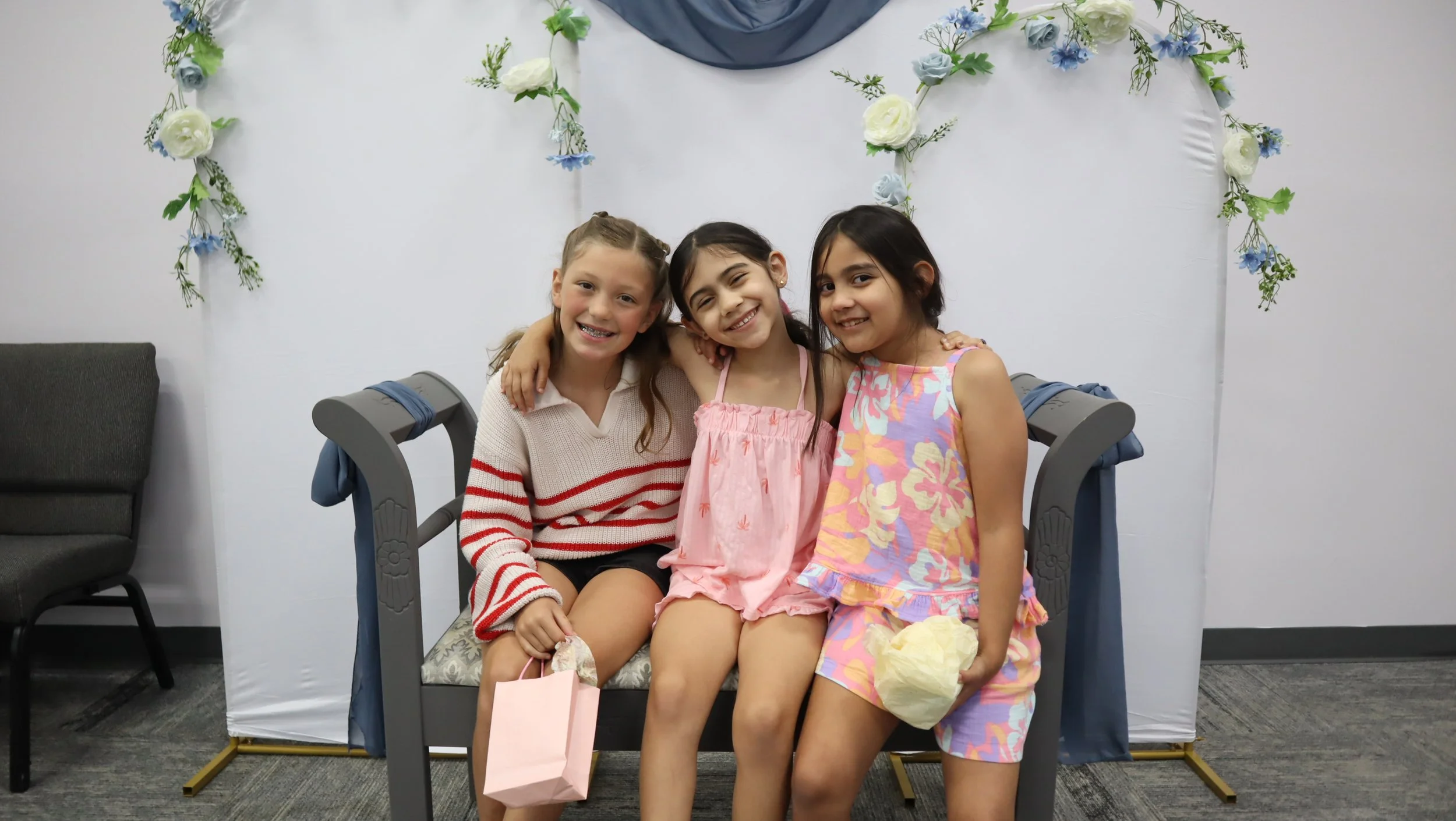 Three girls sitting close together, smiling, with one girl having her arm around another, in a room with floral decorations on the wall behind them.