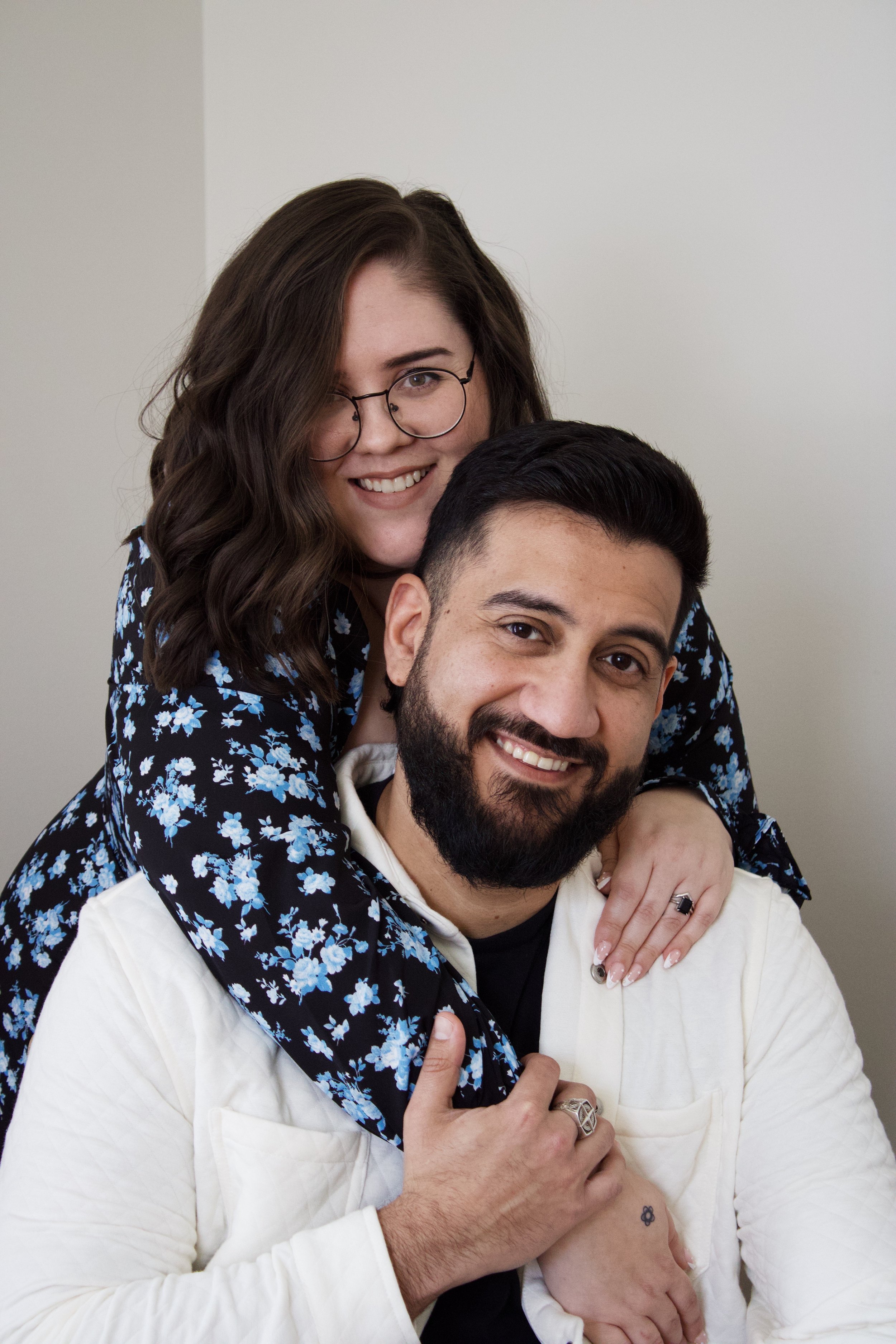 A woman with glasses and wavy brown hair smiling, hugging a man with a beard and short dark hair, both wearing floral patterned and plain white clothing respectively, against a plain light-colored background.