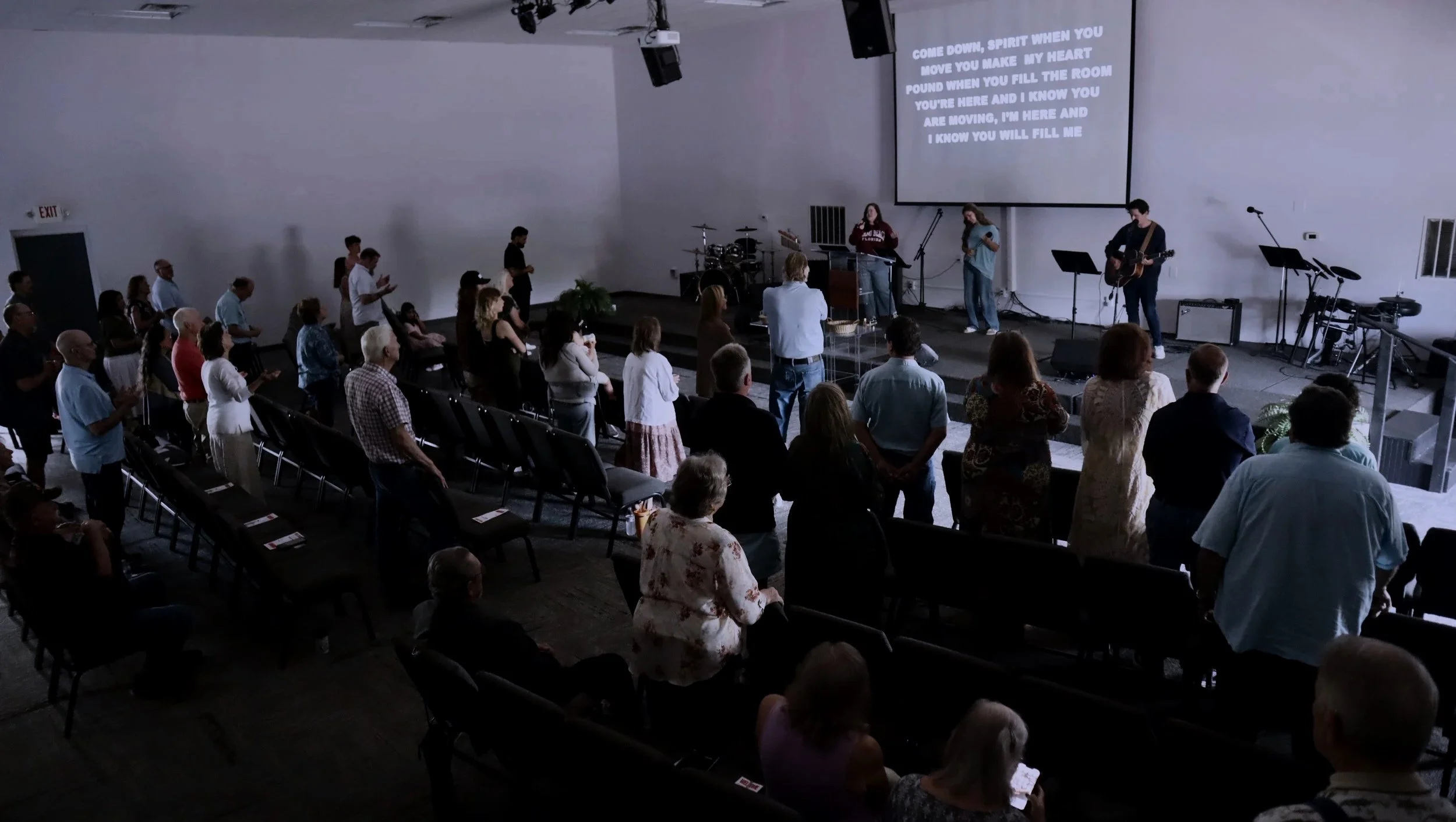 A group of people gathered in a church auditorium, standing and facing the stage where three musicians are worshipping Jesus. The stage has a large screen displaying lyrics for a song, and there is musical equipment including drums and microphones.