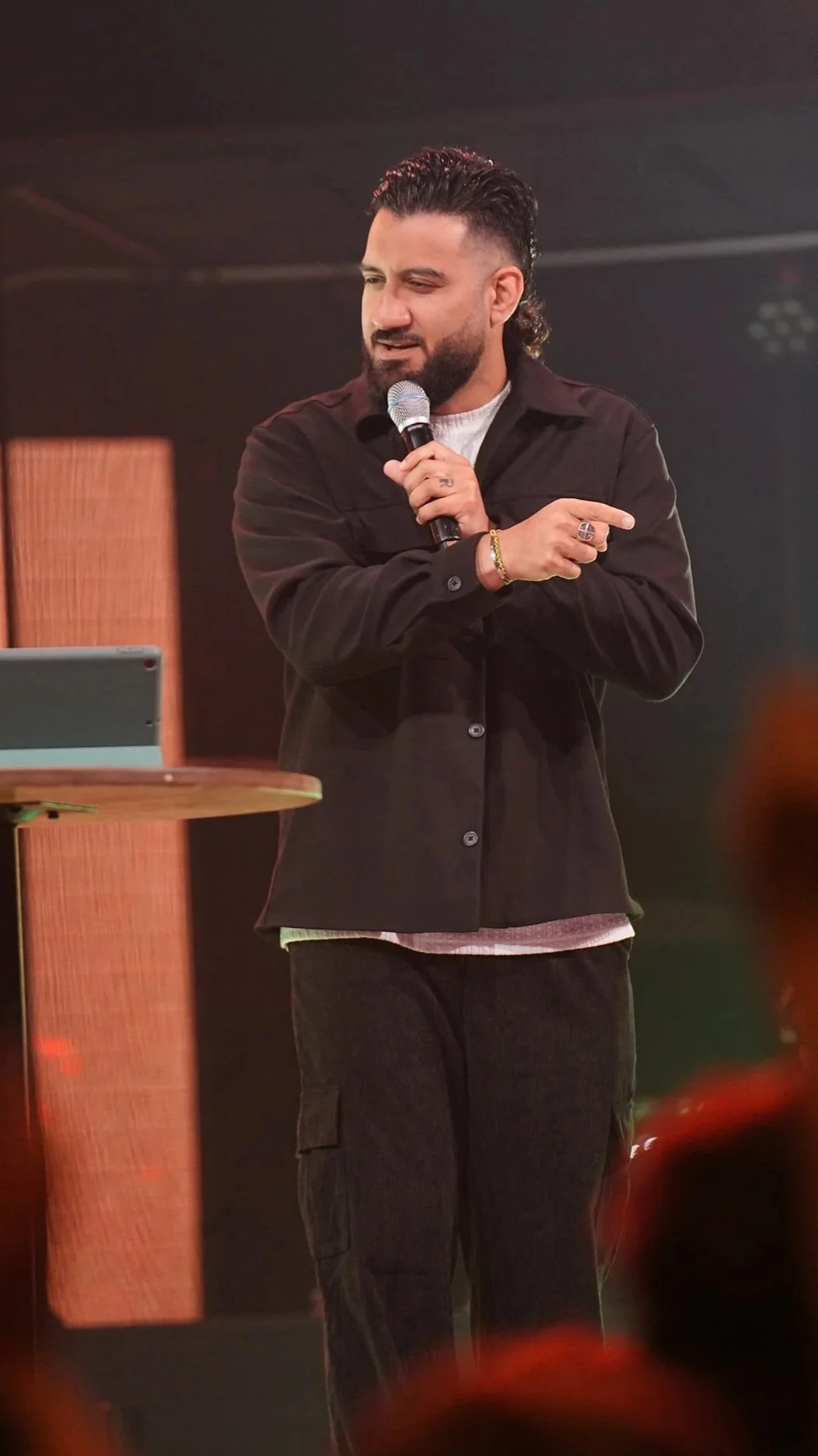 A man with dark hair and a beard wearing a black jacket and cargo pants, holding a microphone, speaking or performing on stage.