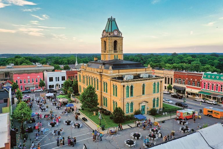Downtown area with a historic courthouse at the center, surrounded by colorful shops and tables with chairs for outdoor dining on a lively street during daytime.