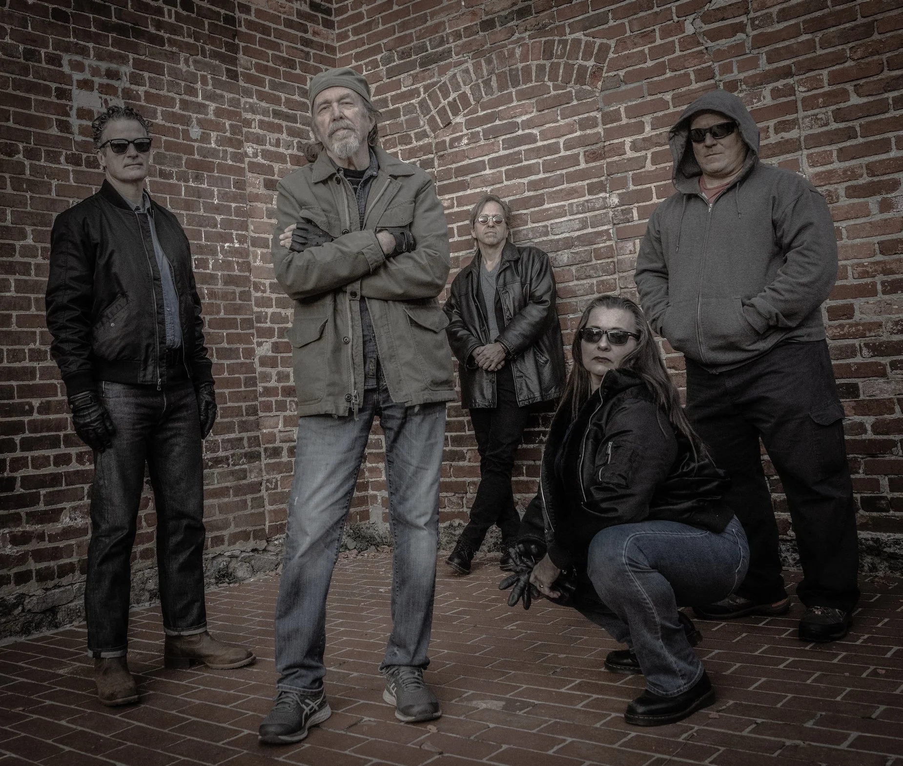 Five people standing in front of a brick wall, wearing dark sunglasses and casual clothes, with one person kneeling.