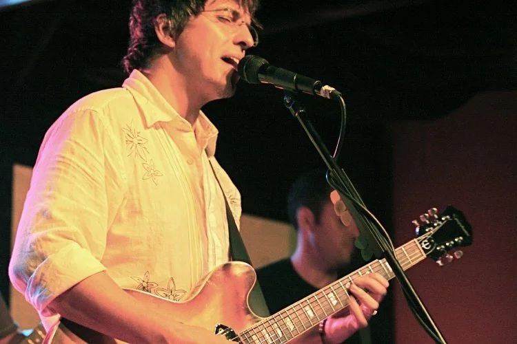 A man singing into a microphone while playing an electric guitar, with another band member in the background.