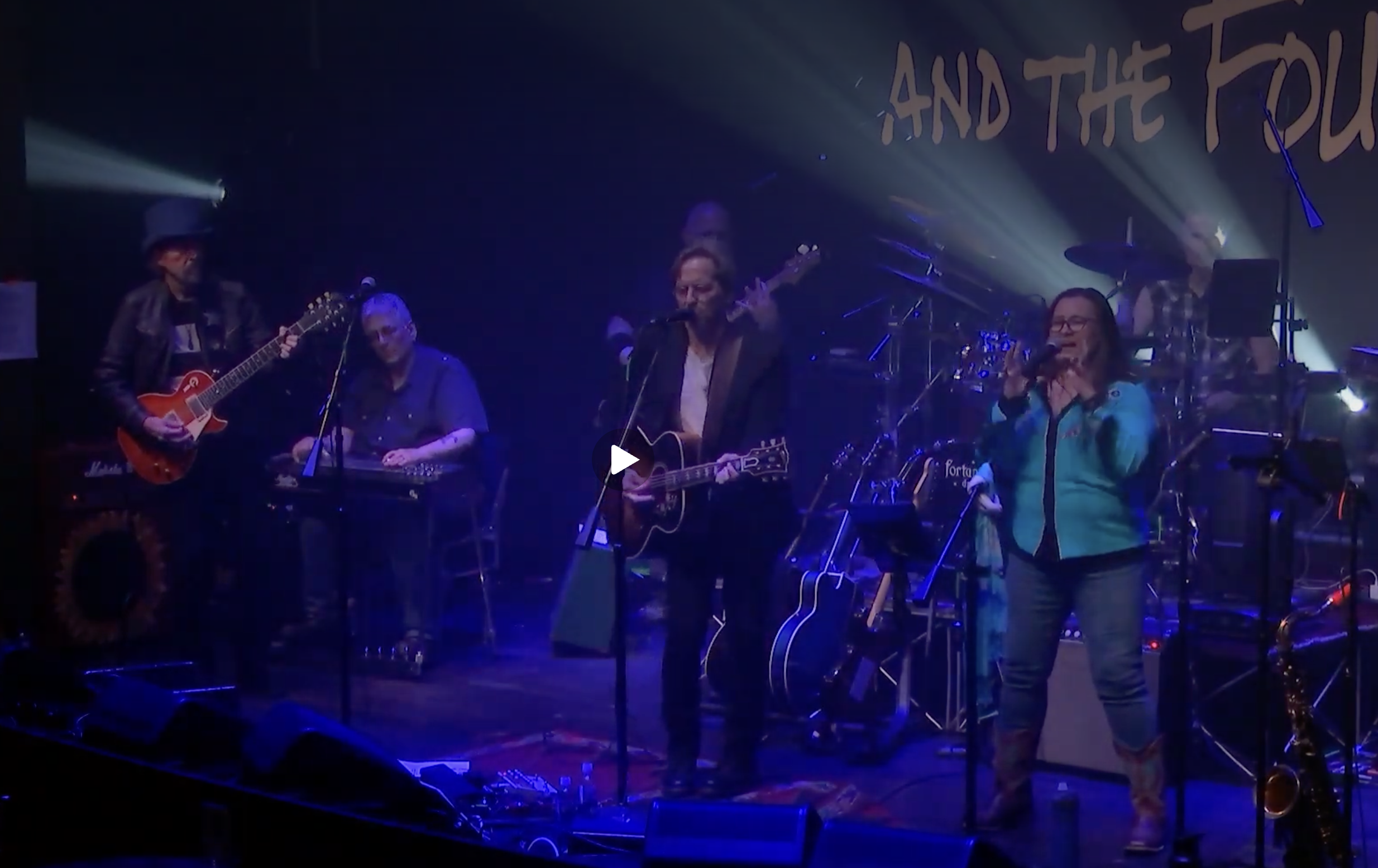 Band performing on stage with multiple musicians, including guitarists and a vocalist, under colorful stage lighting. A large sign in the background reads 'AND THE...'.