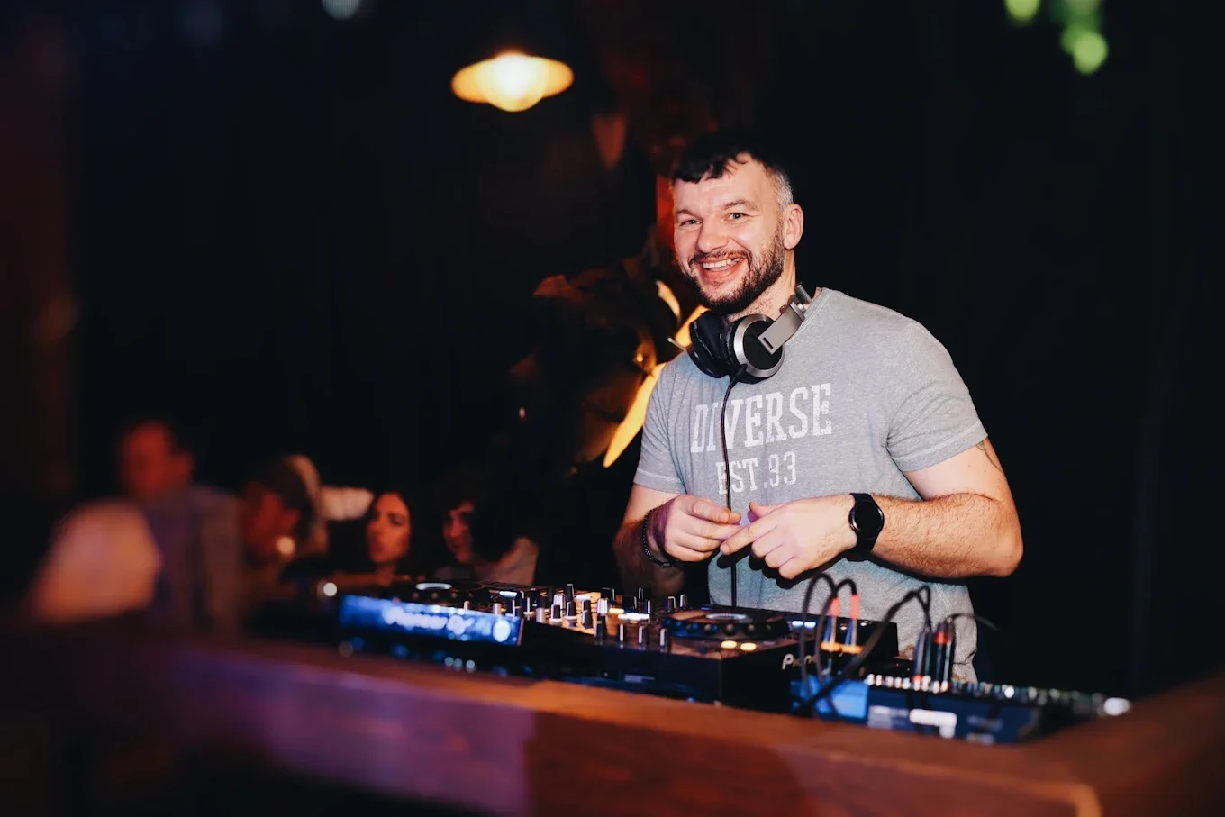 „DJ ze słuchawkami na szyi, uśmiechnięty podczas obsługi sprzętu DJ-skiego w ciemnym klubie, z publicznością w tle.