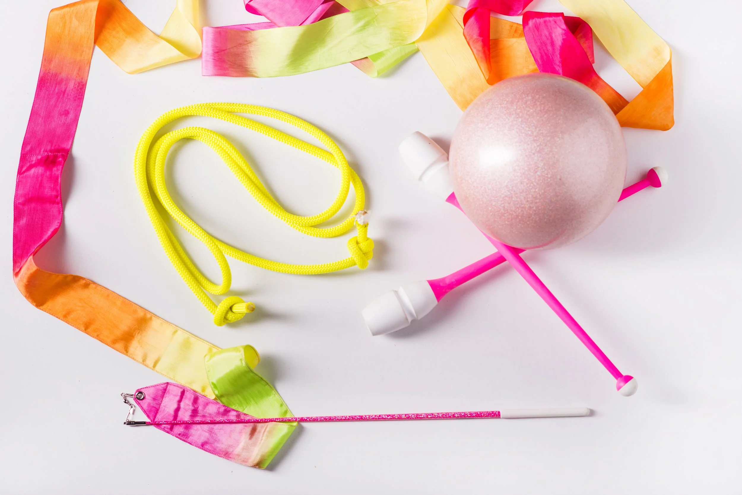 Colorful party decorations and accessories, including pink, yellow, and orange ribbons, a pink ball ornament, yellow string, a pink magic wand, pink and white pushpins, and a pink flag, arranged on a white surface.