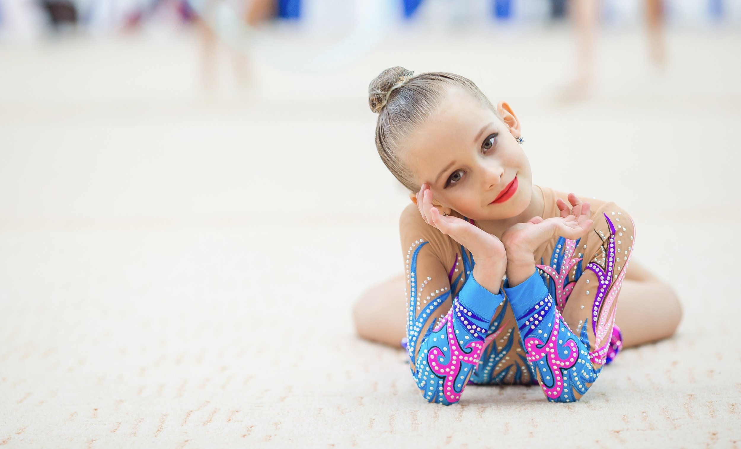 A young girl in a colorful gymnastics leotard with rhinestones, lying on her side on a gymnasium floor, resting her head on her hand, with a focused expression.