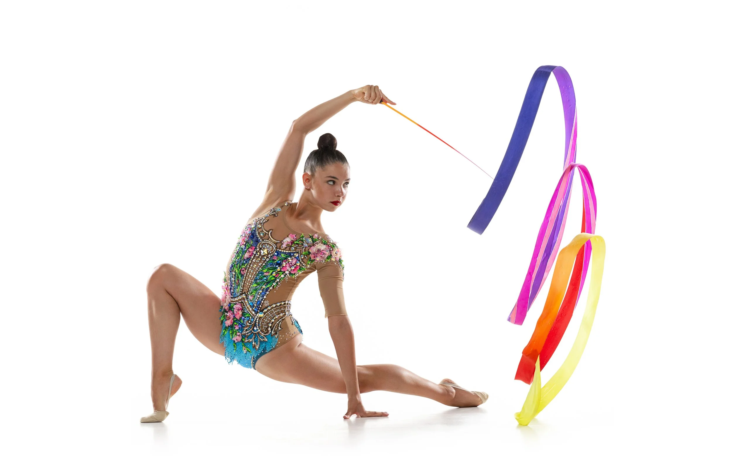 A female gymnast balancing in a pose with one knee on the ground and one leg extended, wearing a colorful, decorated leotard, against a plain white background, with multicolored ribbons in motion nearby.