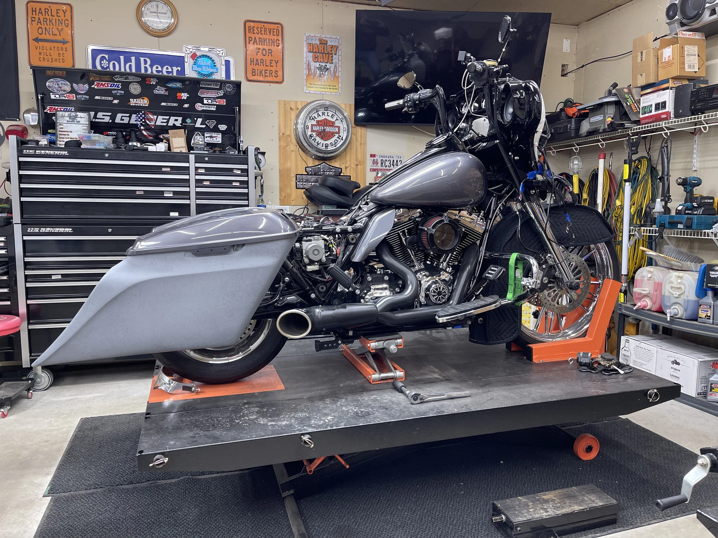 A motorcycle is being repaired or built in a garage. It is mounted on a stand, with tools and part organizers surrounding it. The garage has signs on the wall, a flat-screen TV, and various equipment and supplies on shelves.