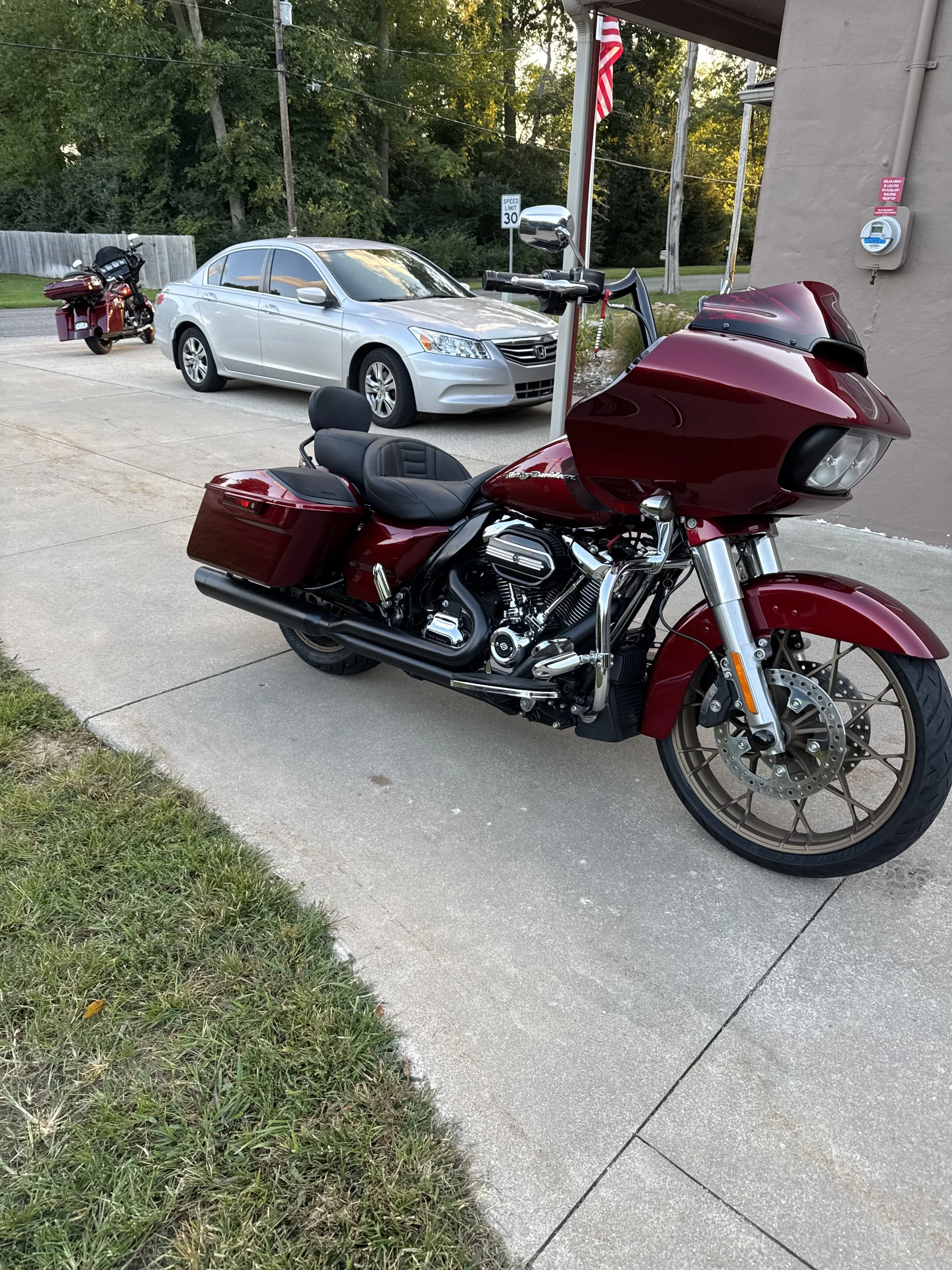 Motorcycle in foreground with large front fairing, black seat, and burgundy color, parked on concrete sidewalk. Silver sedan and red motorcycle in background, near grass and a wooden fence. Green trees, power lines, and a speed limit sign of 30 mph visible in background.