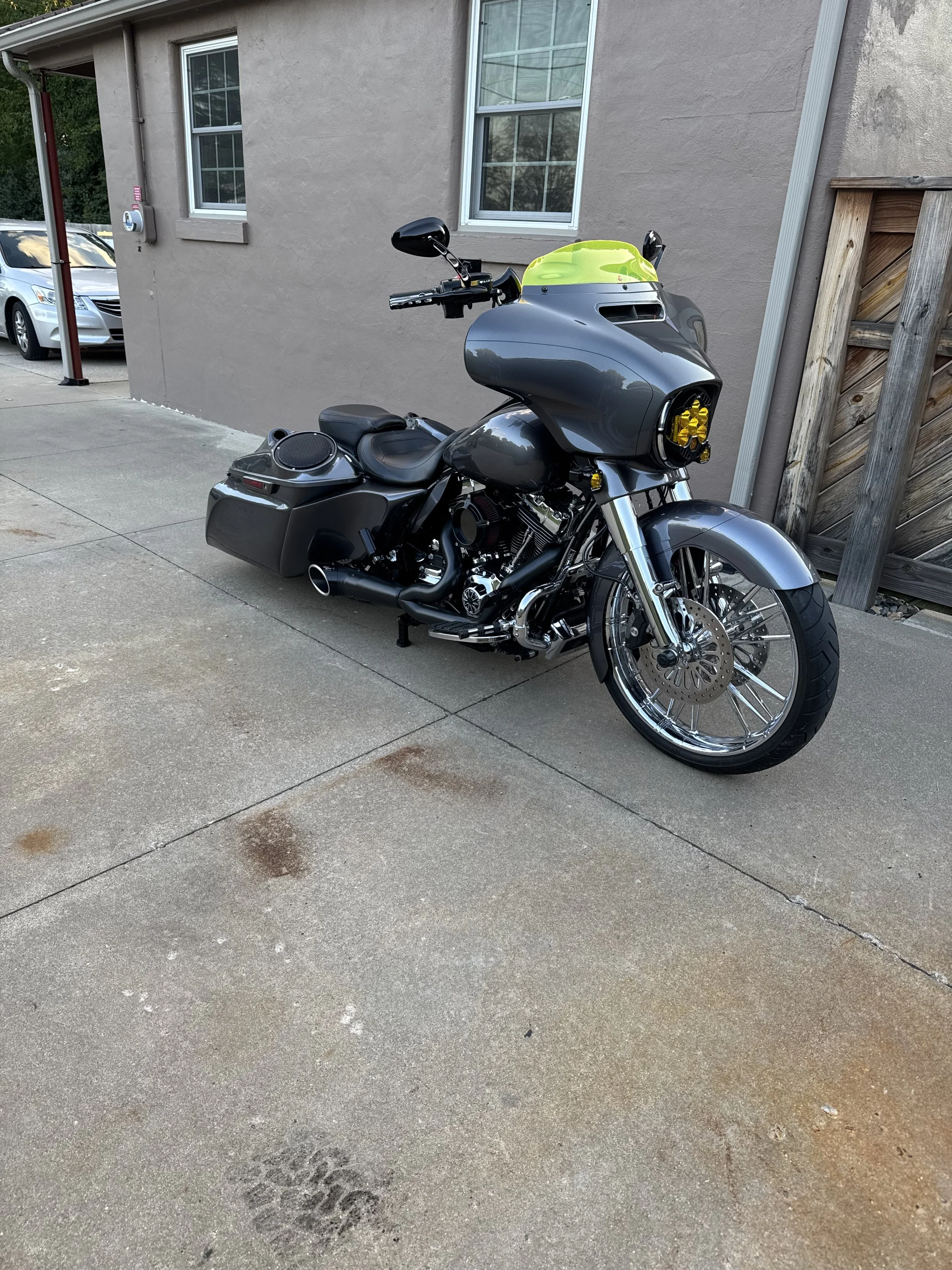 A black motorcycle with yellow headlights and a green helmet on the front, parked on a concrete driveway beside a beige house with two windows.